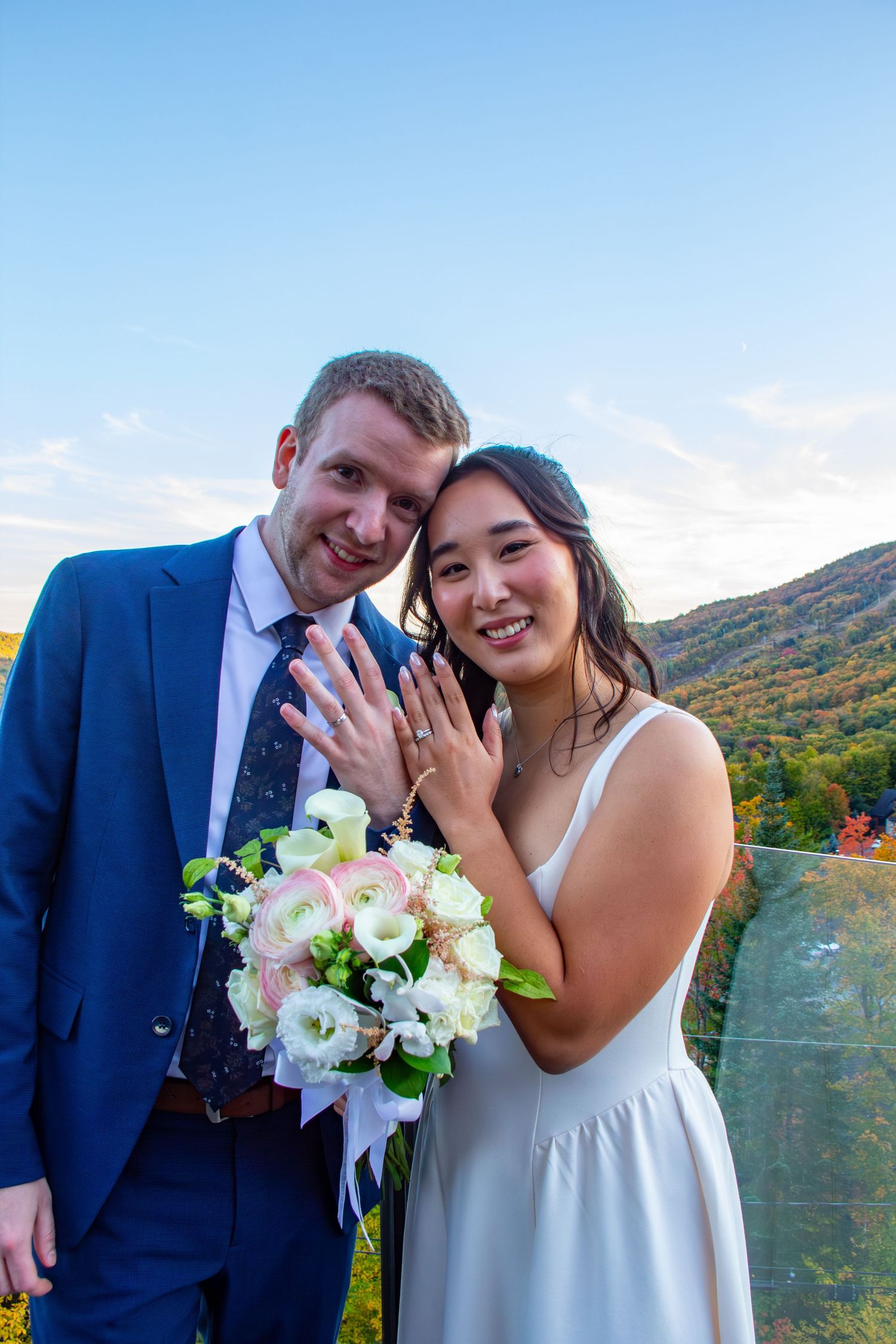 Smiling newlyweds showing wedding rings with autumn mountain scenery in the background.