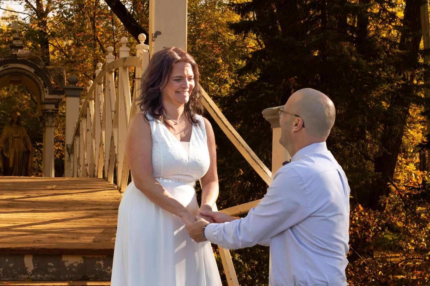 Bride in white dress and groom holding hands on a bridge surrounded by golden autumn foliage.