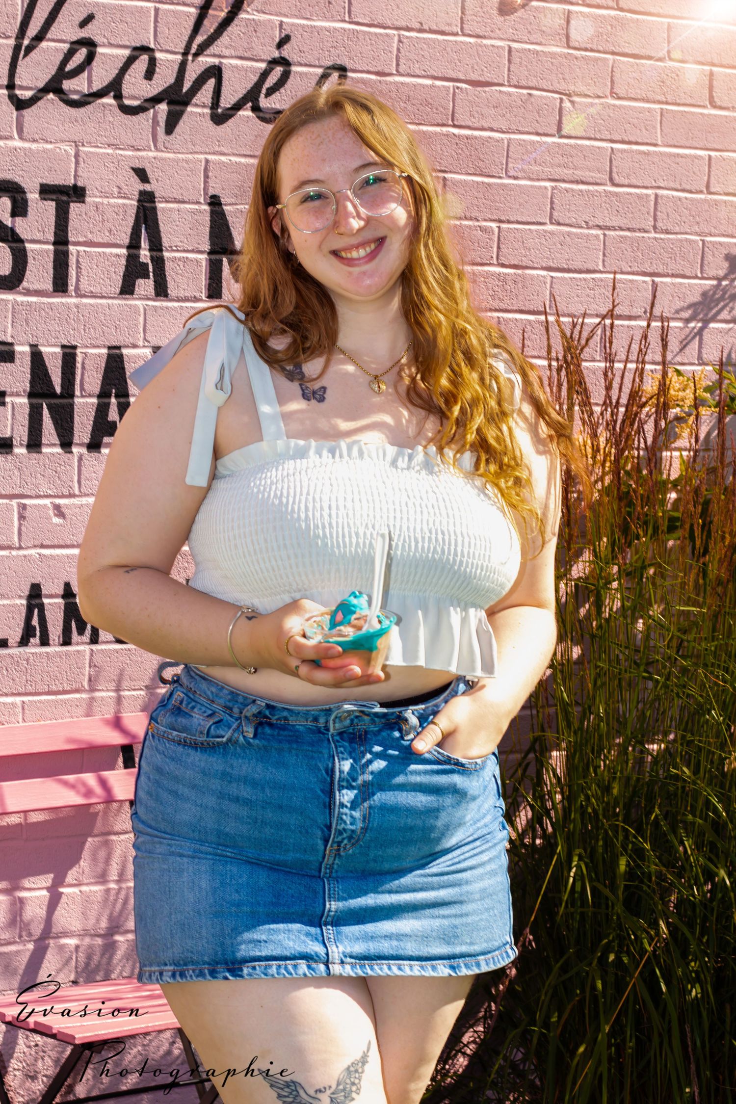 Jeune femme souriante tenant une crème glacée colorée devant un mur rose à l’extérieur d’un commerce. La fille tablée , Cahrlesbourg