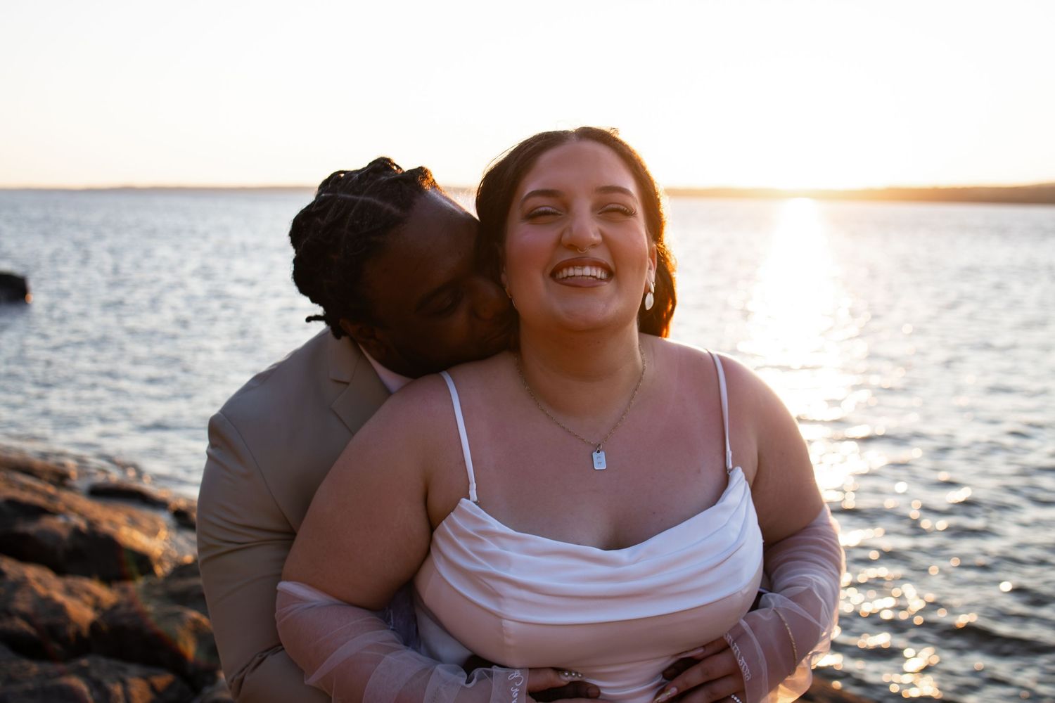 Couple embracing at sunset by rocky shoreline, woman smiling joyfully as man kisses her cheek.