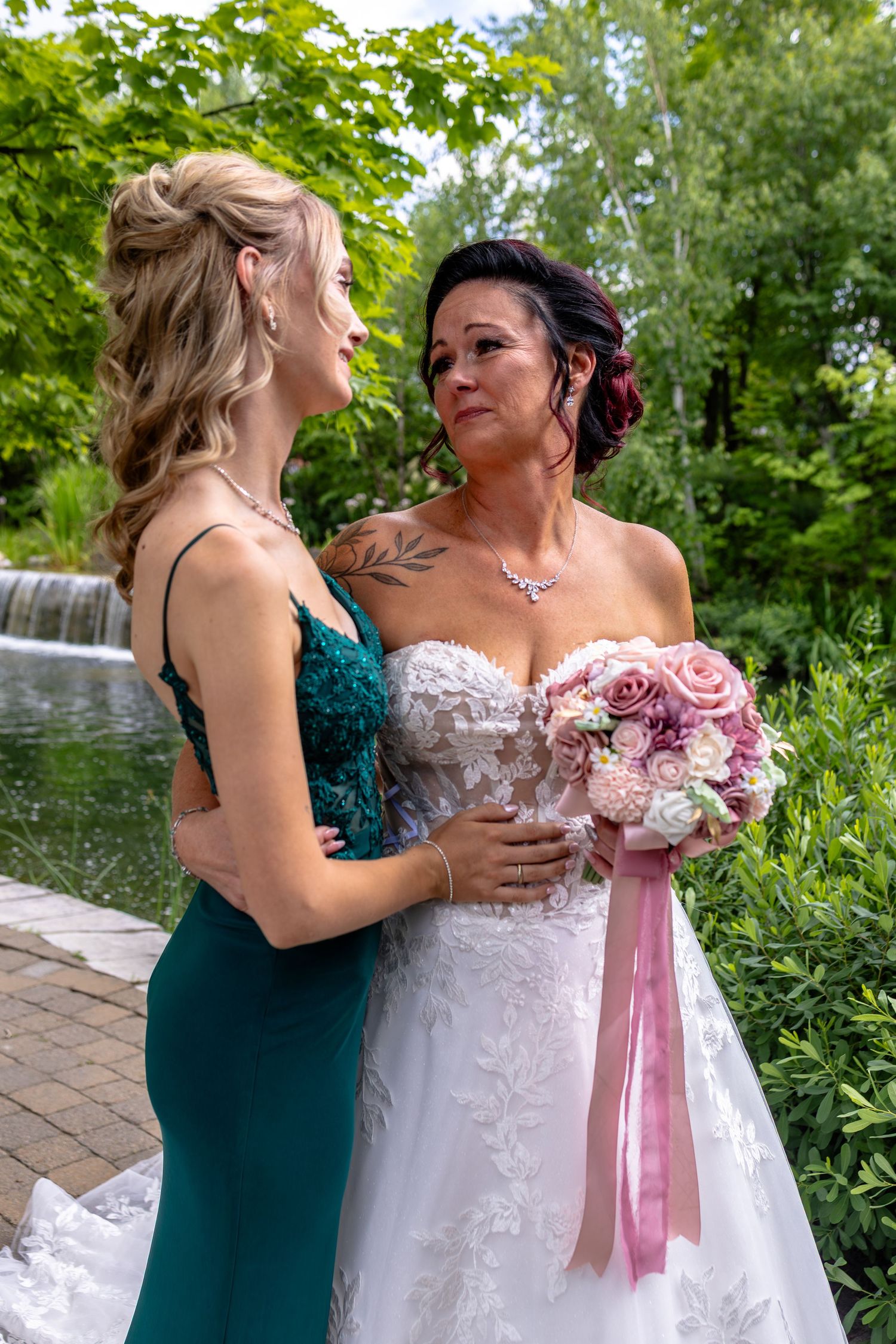 Bride in white lace gown holding pink bouquet stands with bridesmaid in green dress near garden waterfall.
