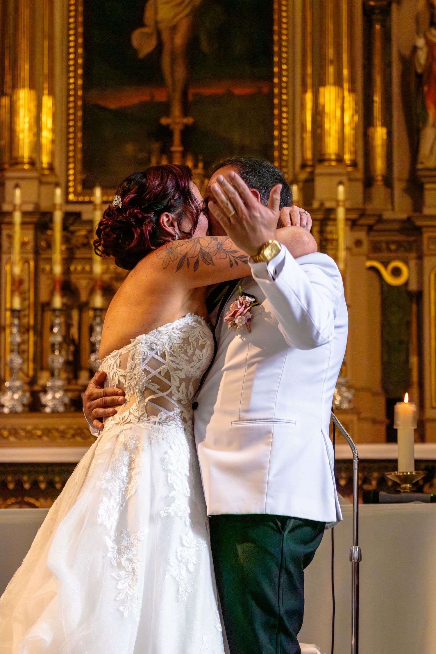 Bride and groom share their first kiss at the altar in a golden church, bride in lace gown, groom in white jacket.