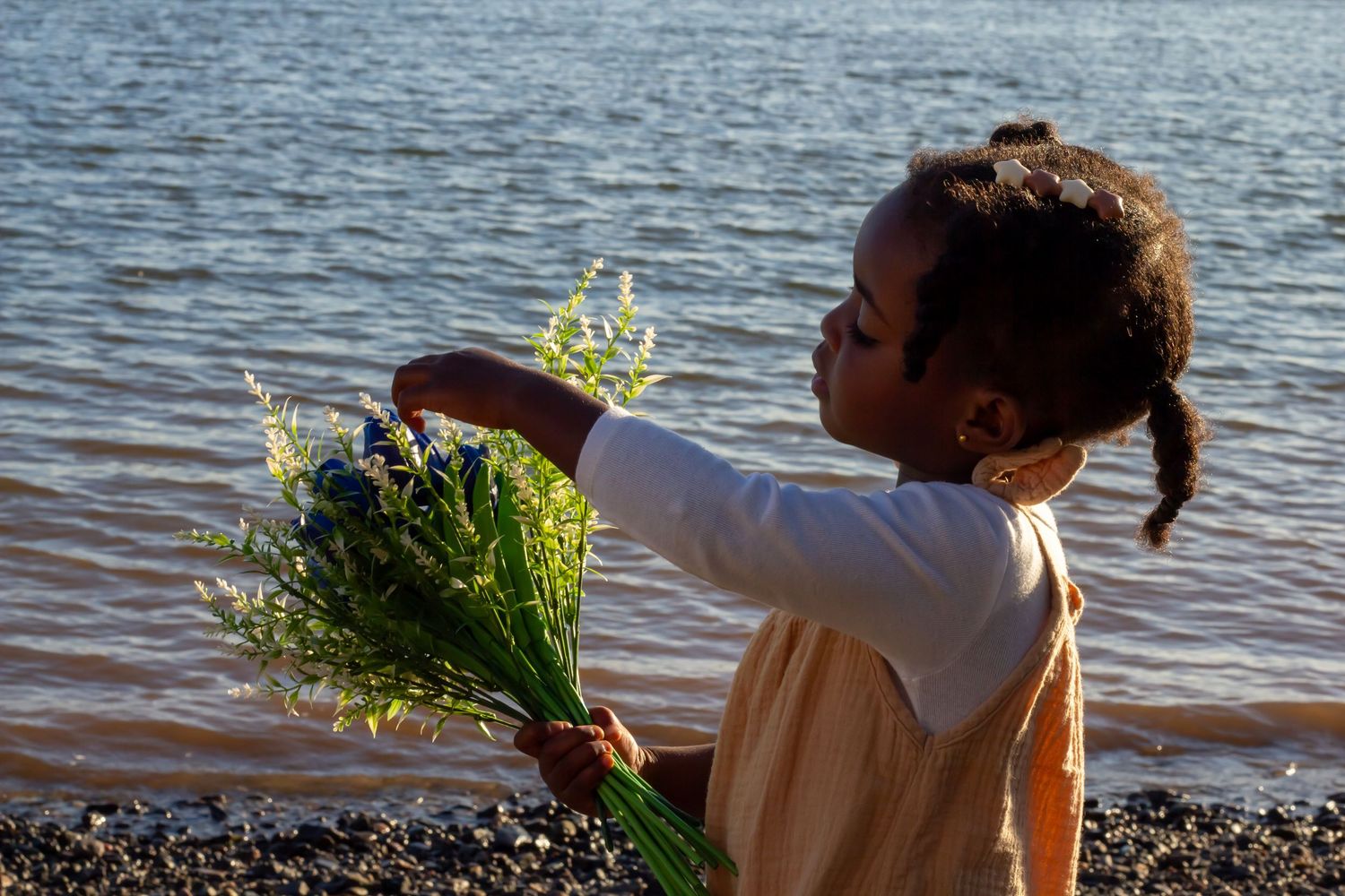 Young Black girl holding wildflower bouquet reaching toward calm river water at sunset.
