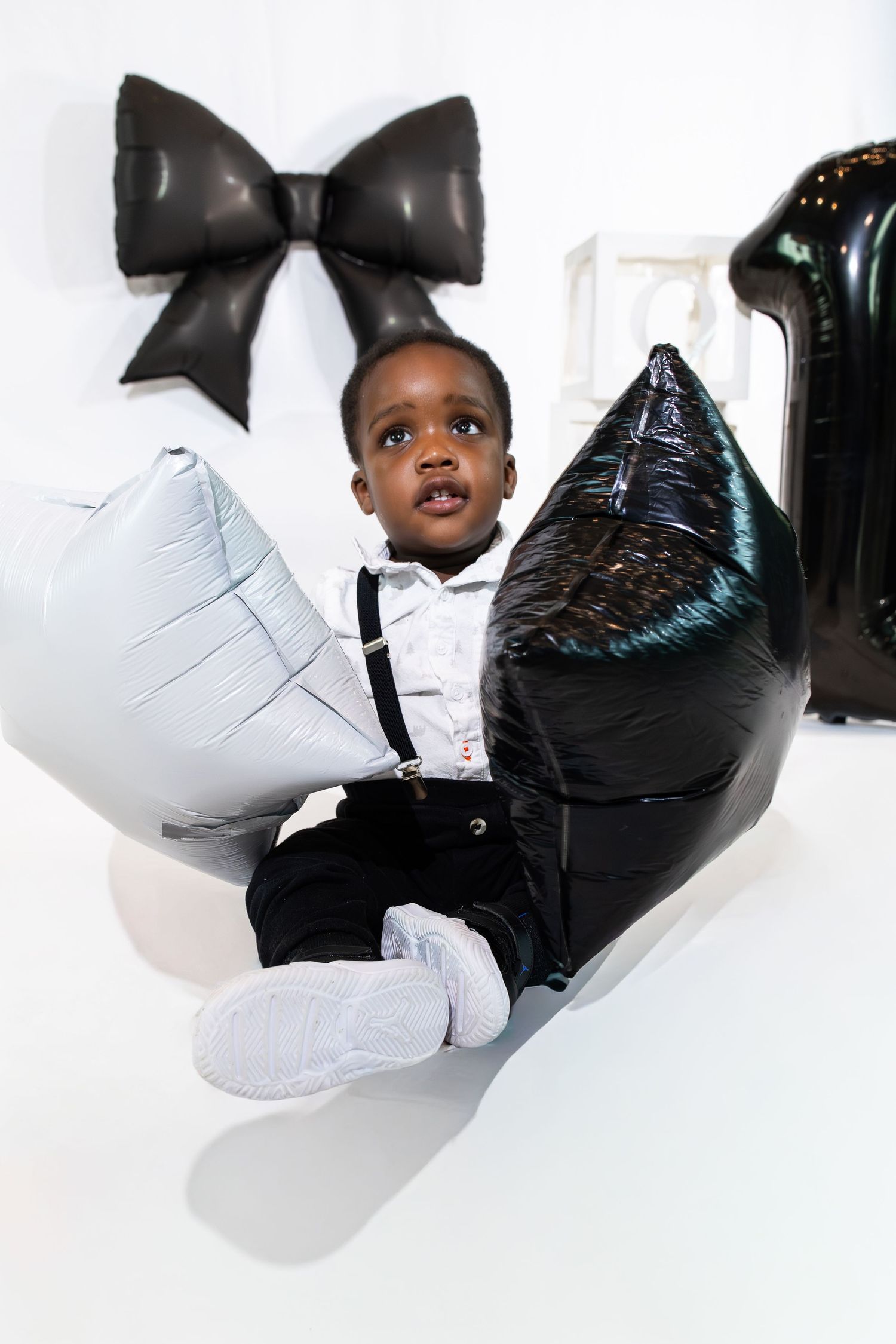 Adorable young boy in suspenders sitting with black and silver star balloons near a bow decoration.