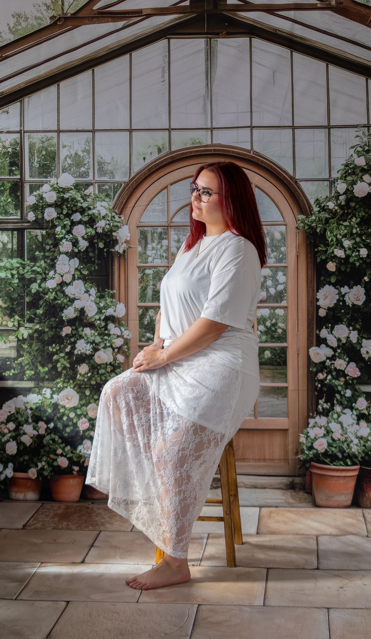 Red-haired woman in white lace dress sits by greenhouse door surrounded by roses.