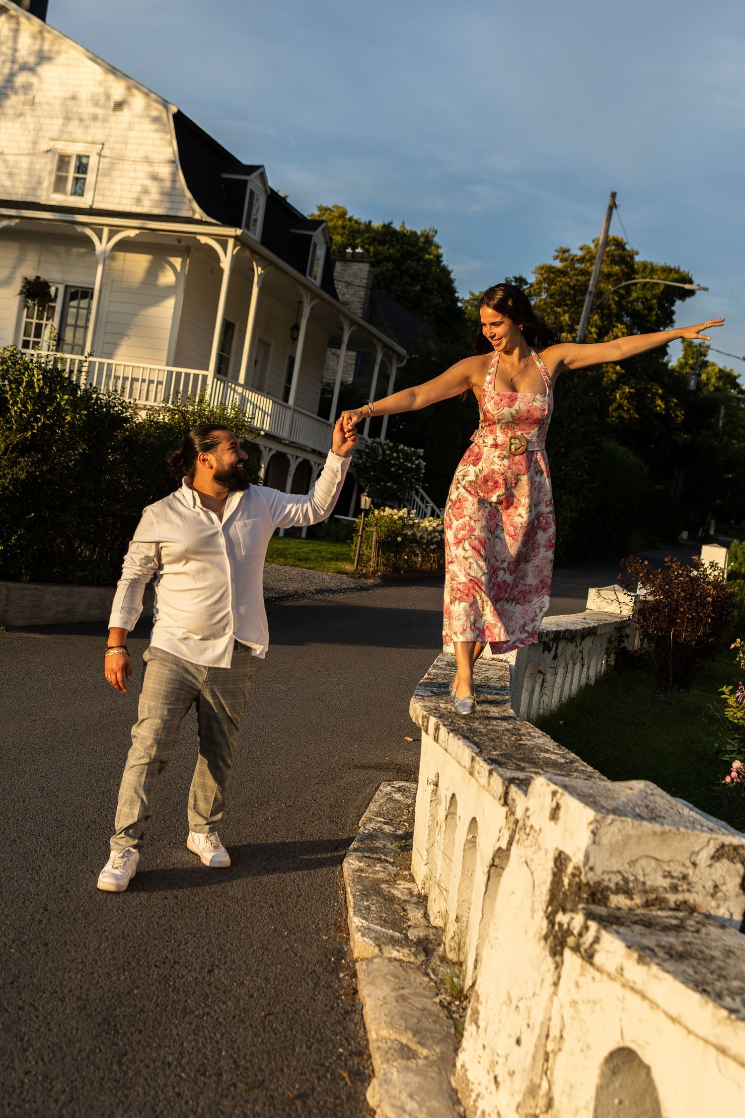 A man in white holds a woman in a floral dress balancing on a stone wall outside a historic white mansion at sunset.