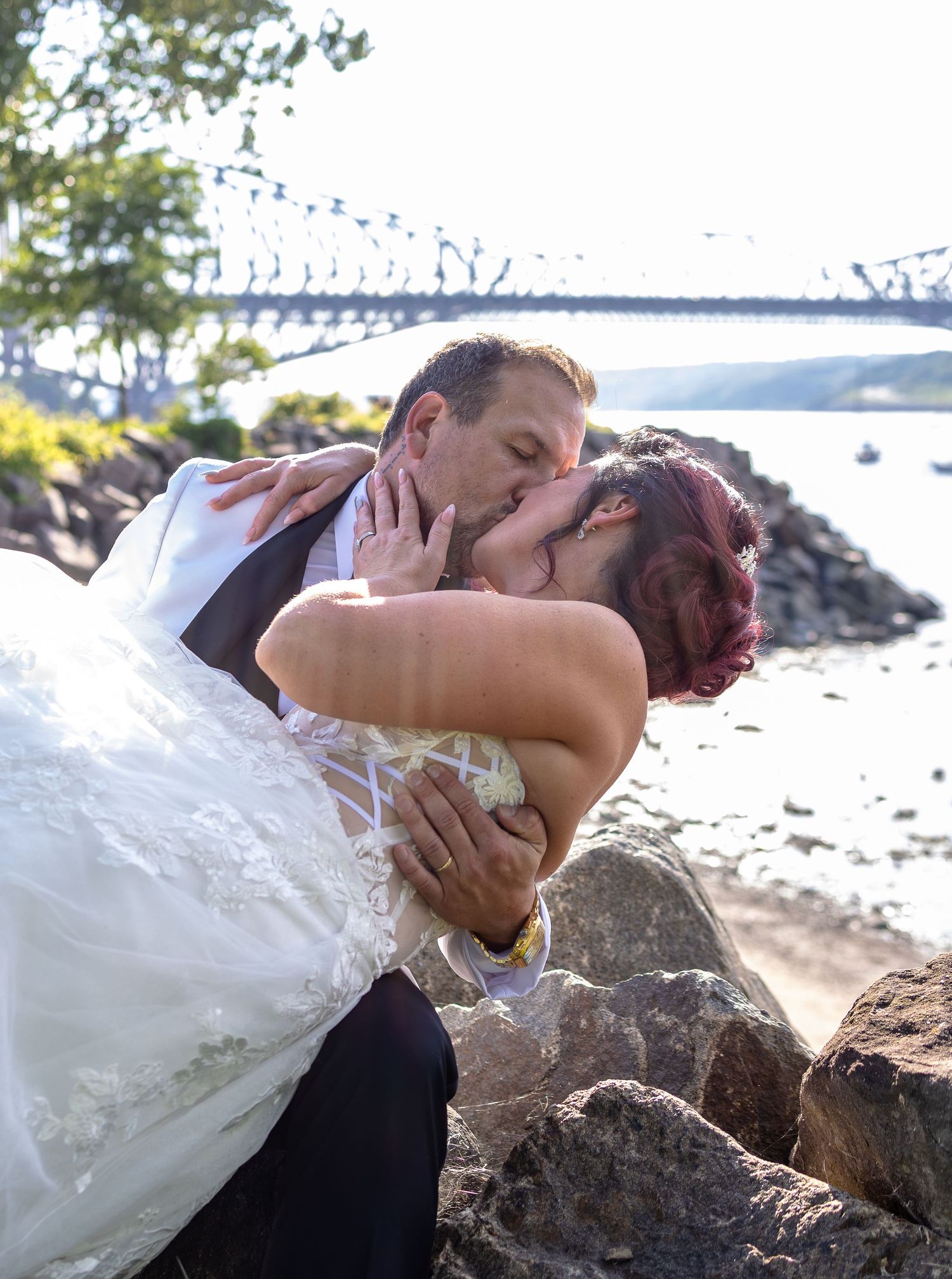Bride and groom sharing a romantic kiss on rocky shoreline with a bridge and river in the background on their wedding day.