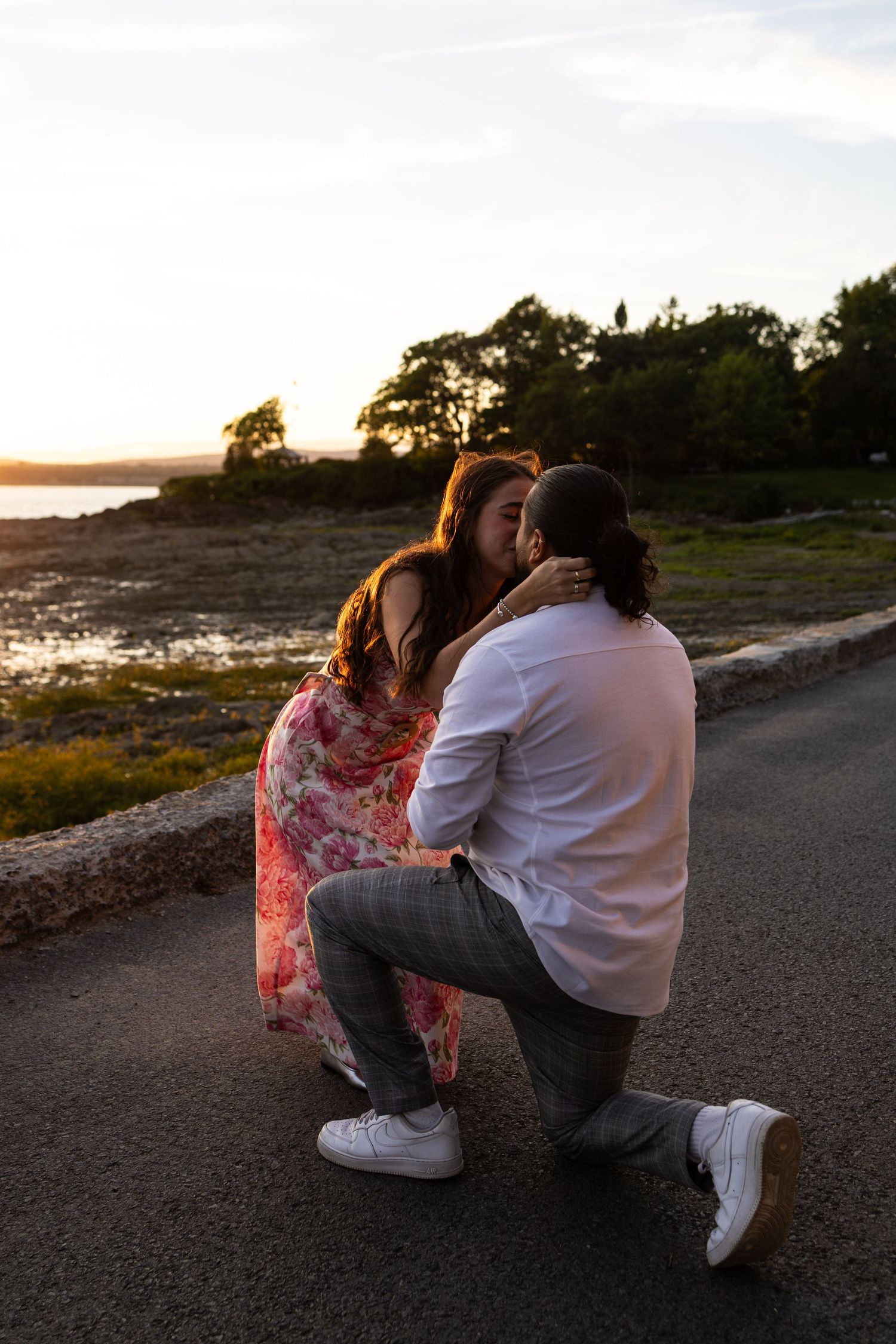 Man on one knee proposing to woman in floral dress at sunset by the ocean.