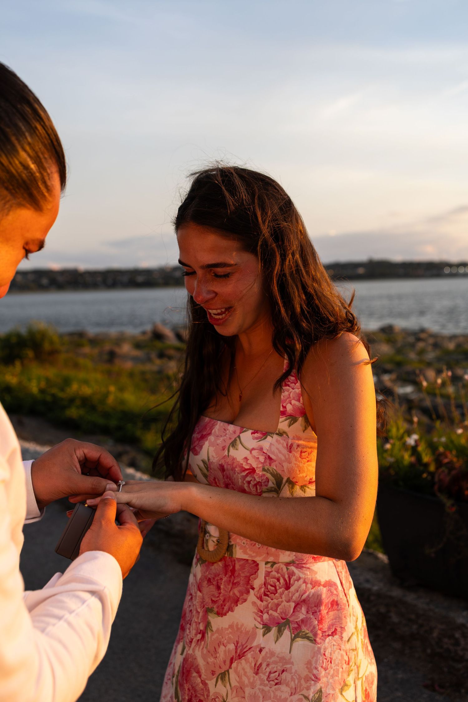 A woman in a floral dress smiles as someone places a ring on her finger at sunset near the water.