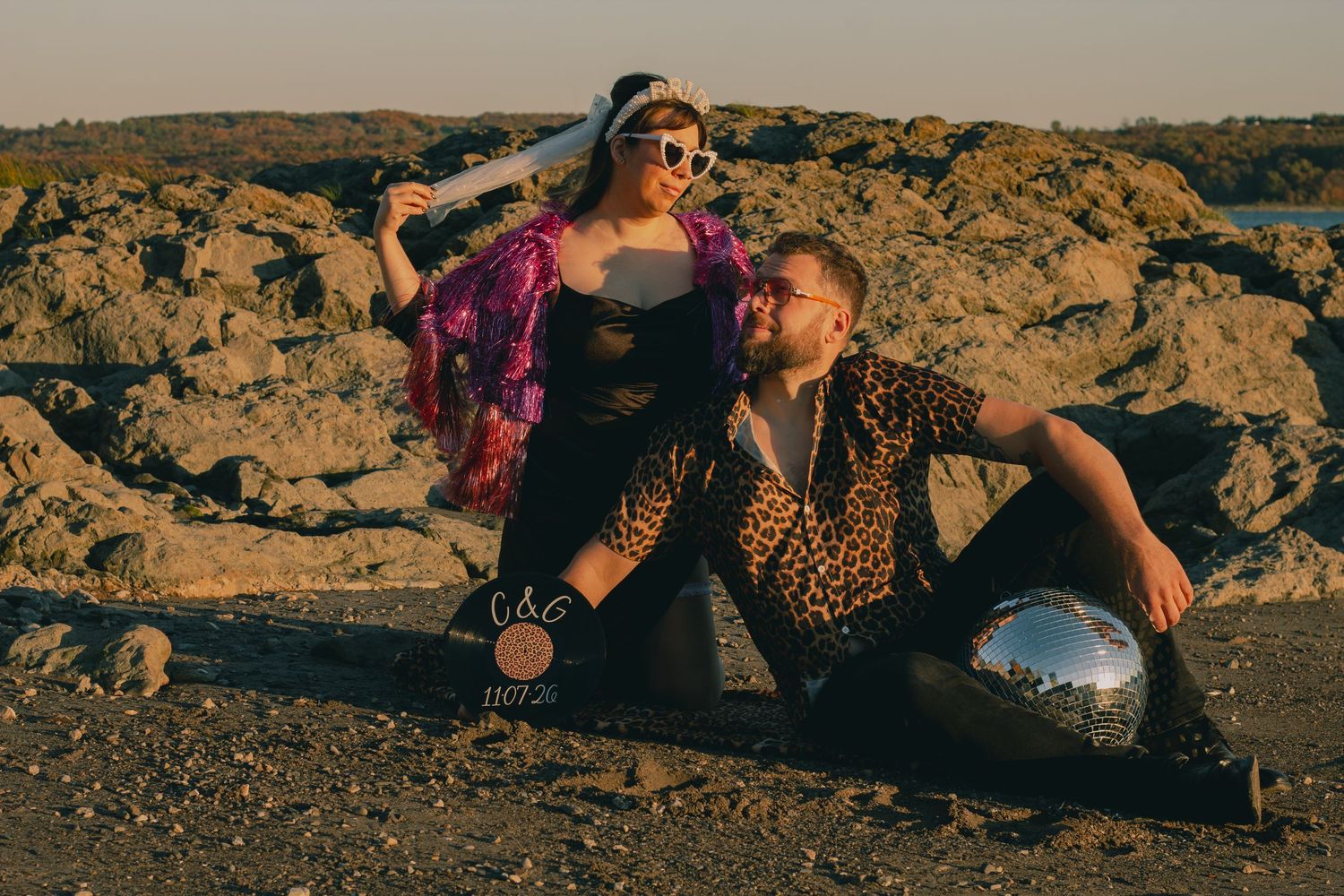 A couple poses outdoors on rocky terrain, woman in purple fur coat and sunglasses, man in leopard shirt with mirror ball.