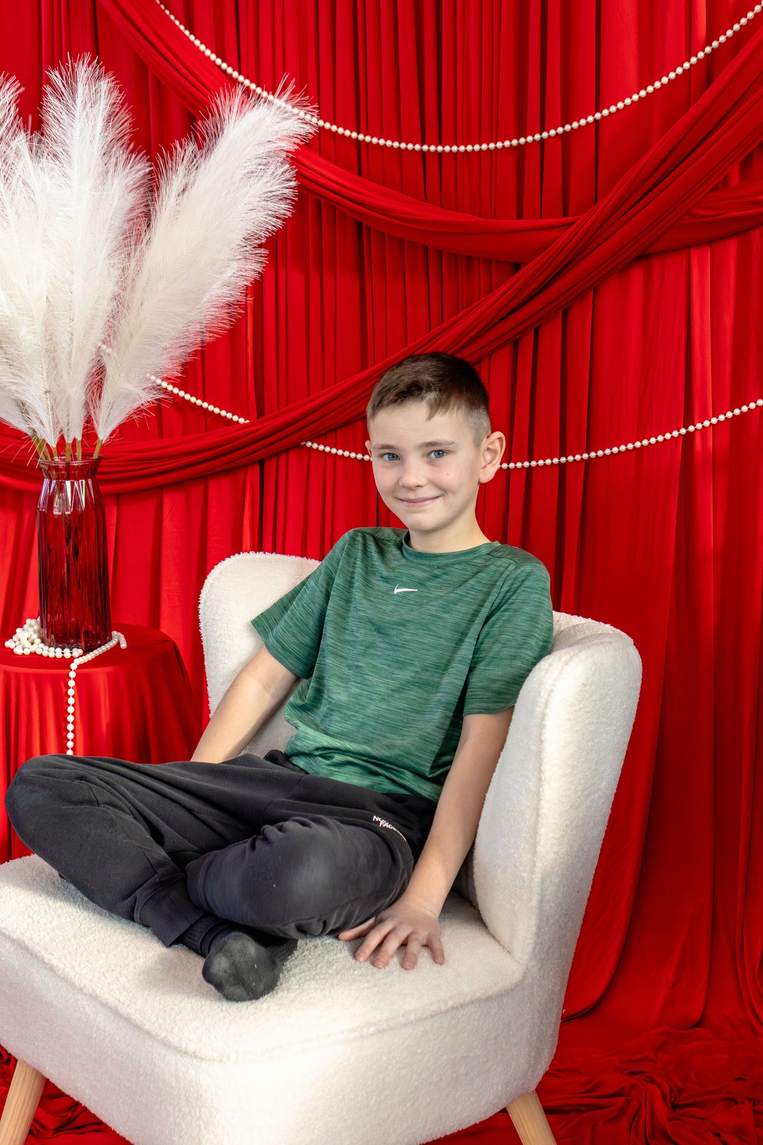 A smiling boy in a green Nike shirt sits cross-legged in a white chair against red curtains with pearl garland decor.