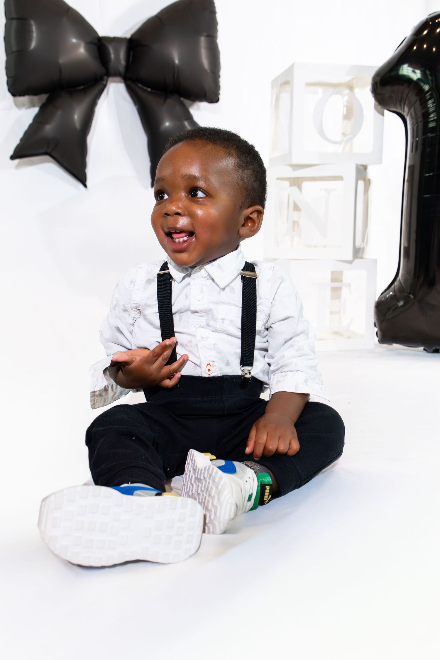 Adorable baby boy in suspenders sits smiling at his first birthday party with a black bow and number 1 balloon.