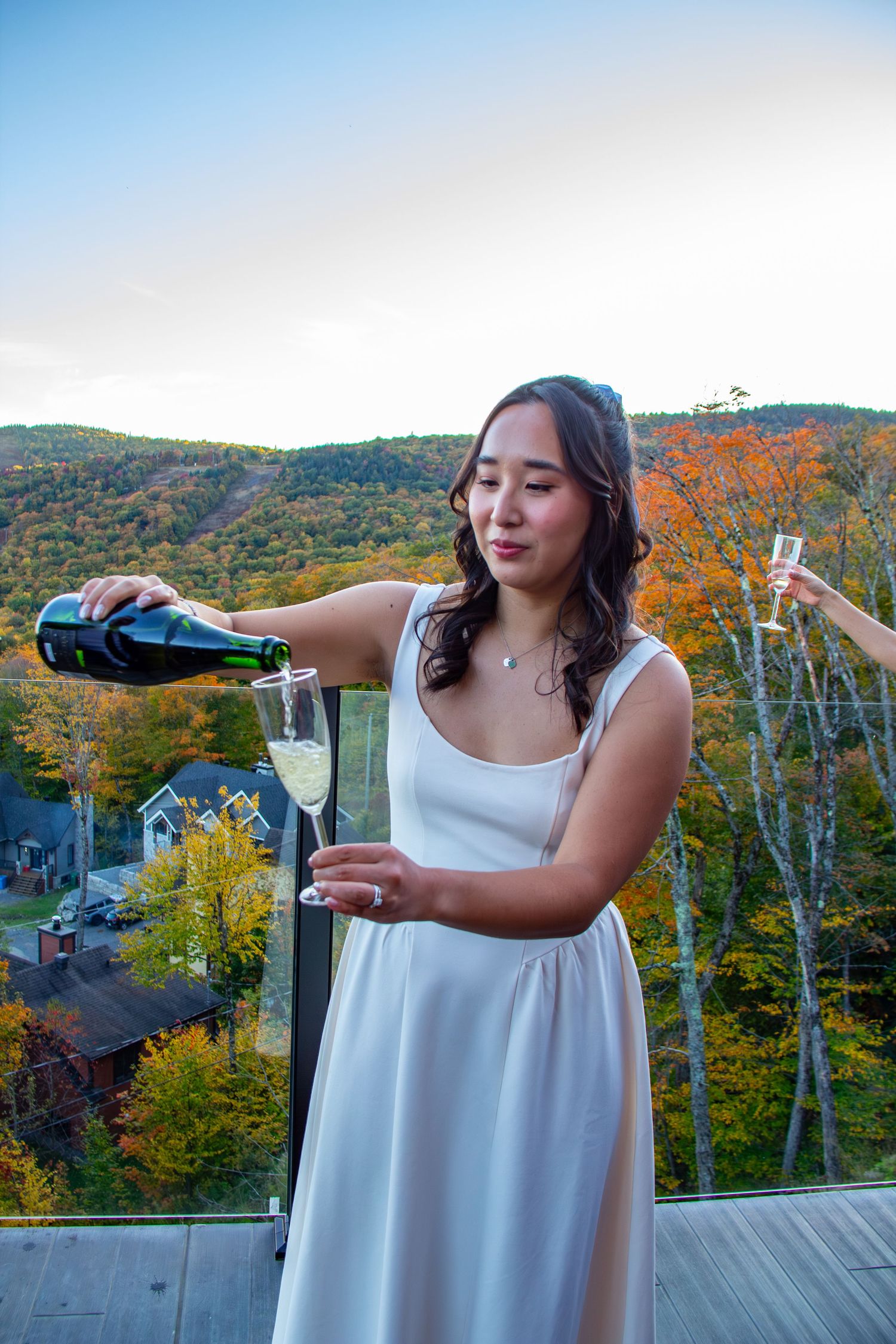 Asian bride in white dress pouring champagne on a balcony with fall mountain scenery.