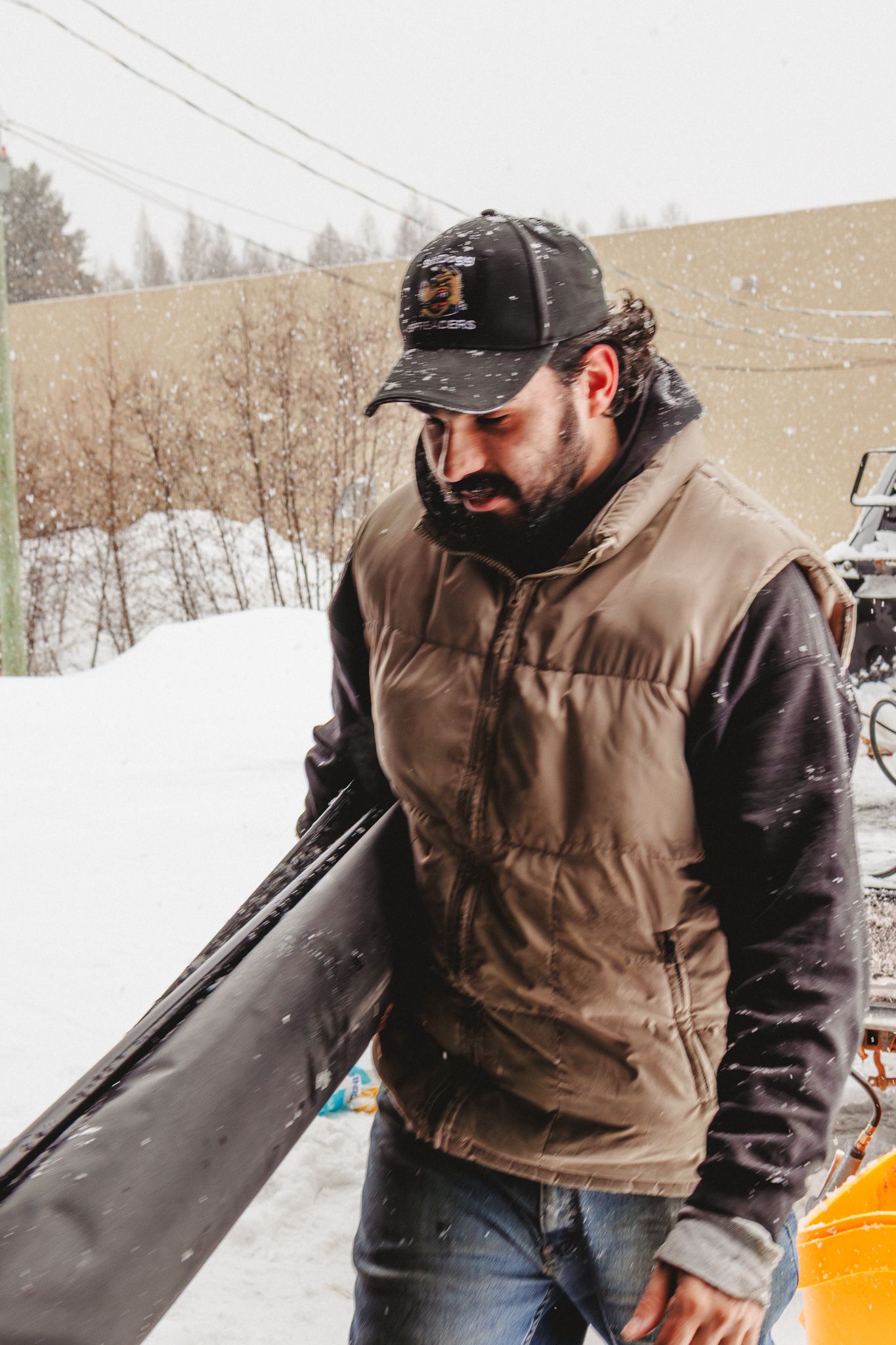 Bearded man in a puffer vest and cap handling equipment outdoors in snowy winter conditions.