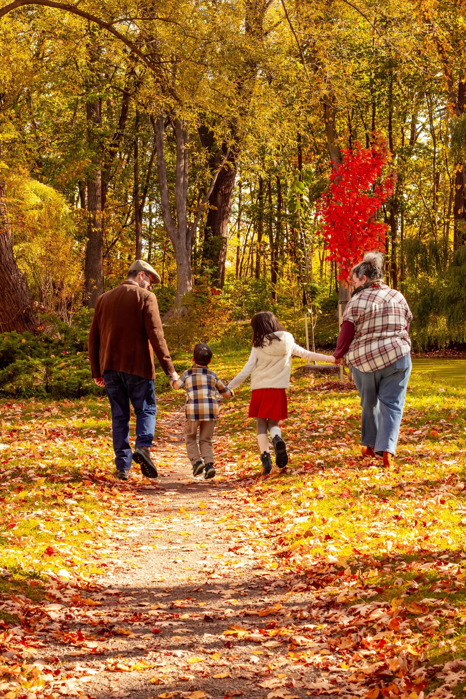 Family of four walking away hand-in-hand along a leaf-covered path through a golden autumn park.