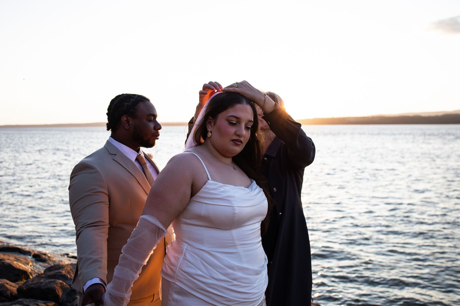 Three people pose by a waterfront at sunset, woman in white dress with colorful hair accessory, man in tan suit behind her.