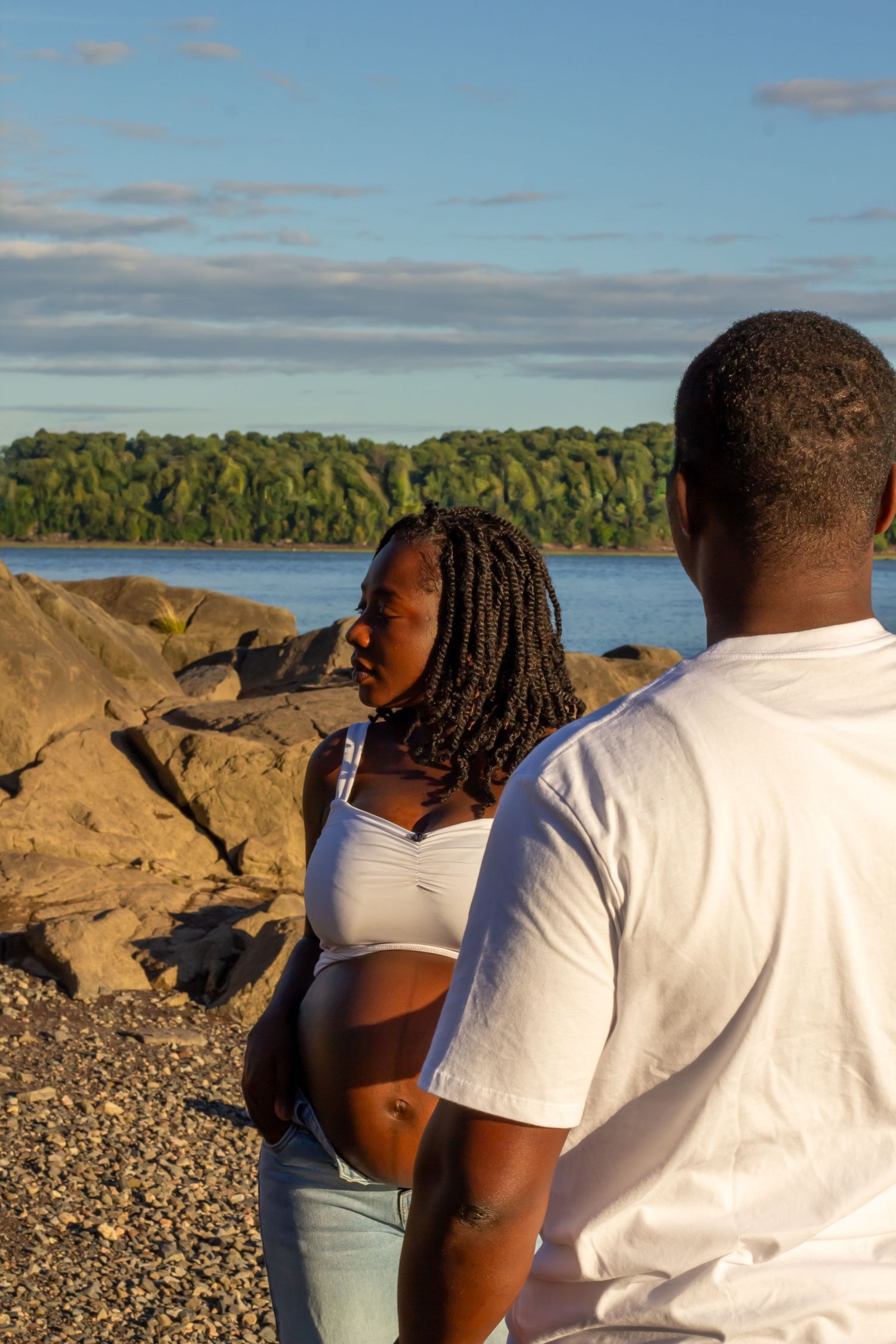Pregnant Black woman in white crop top sharing tender moment with partner by rocky river.