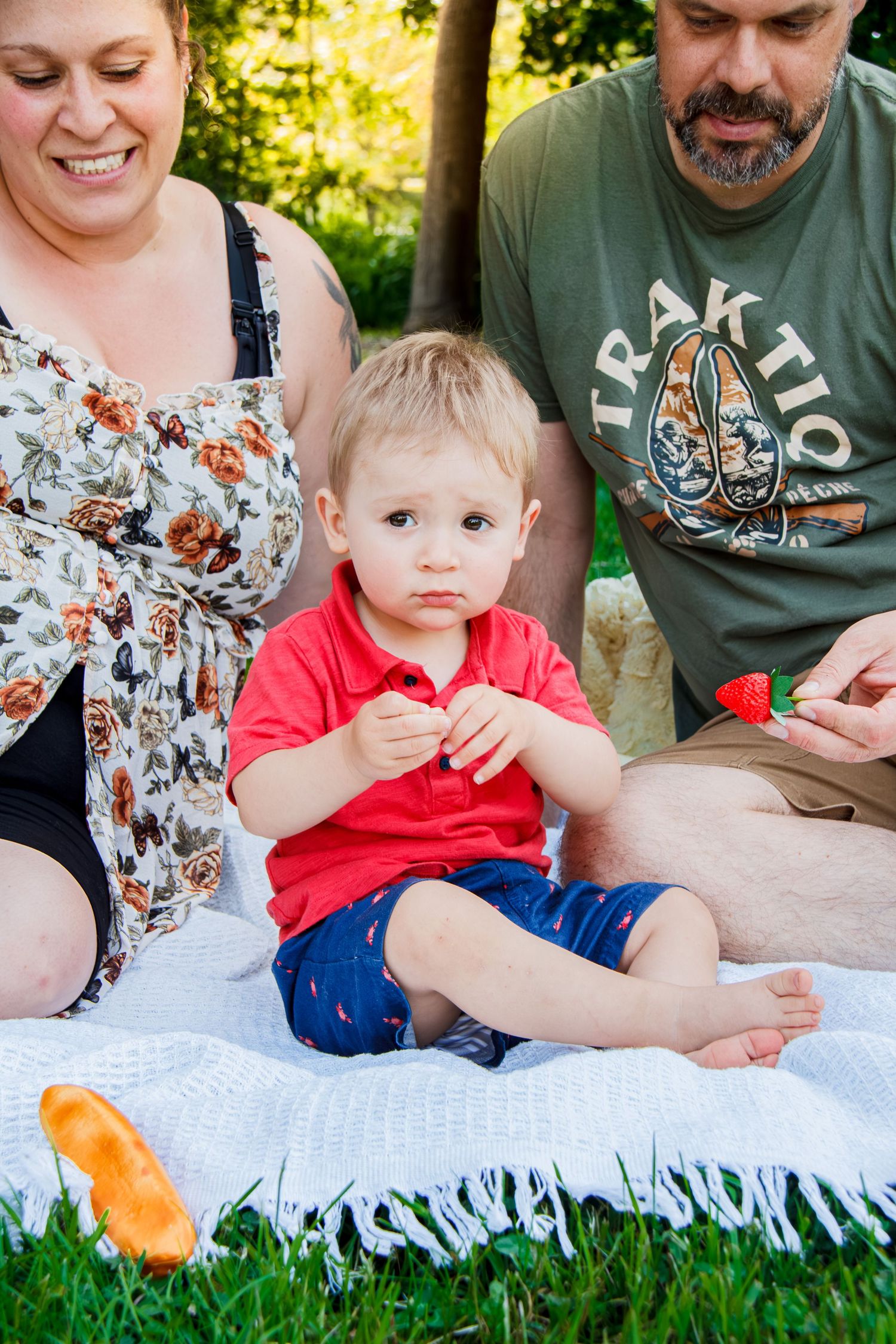 A toddler in a red shirt sits on a blanket holding a strawberry with his parents in a sunny park.