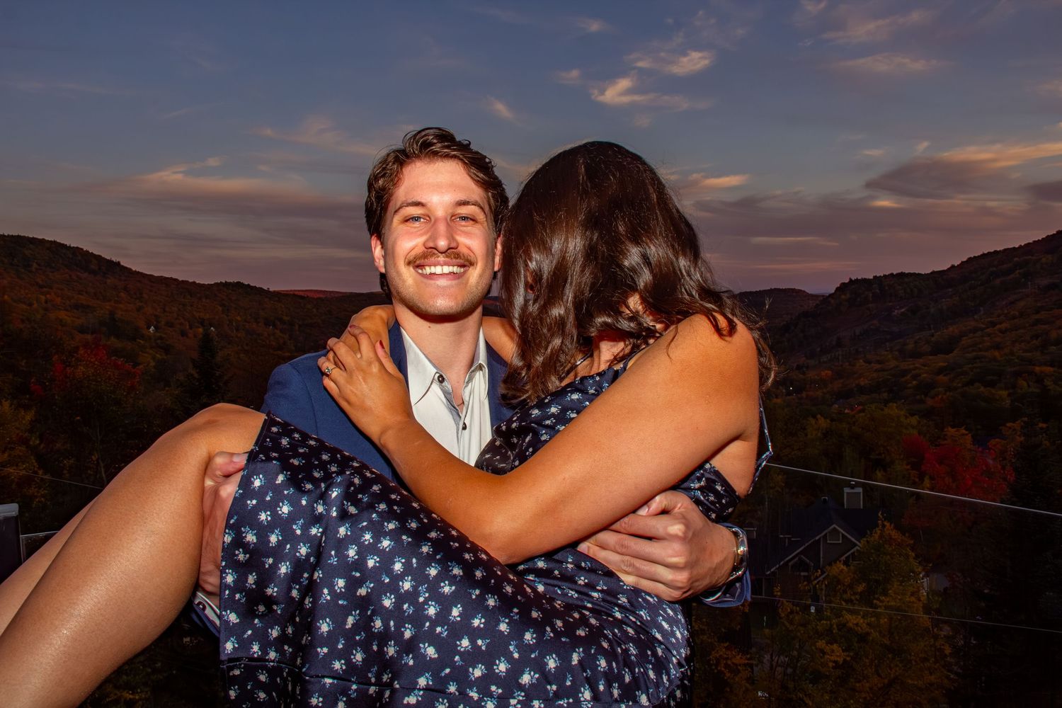 Smiling groom embraces bride in floral dress against a stunning autumn mountain sunset backdrop.