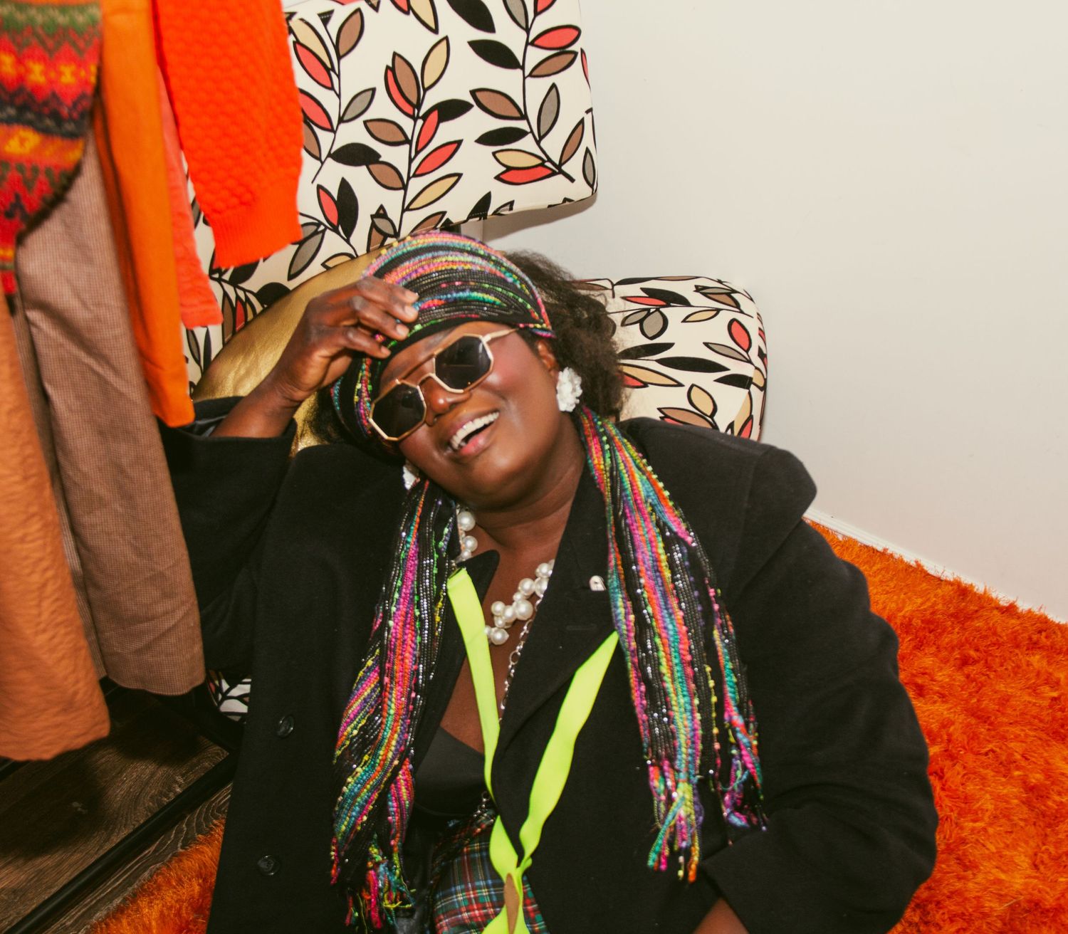 Joyful woman with colorful braids and sunglasses laughing while posing in a vibrant orange room.