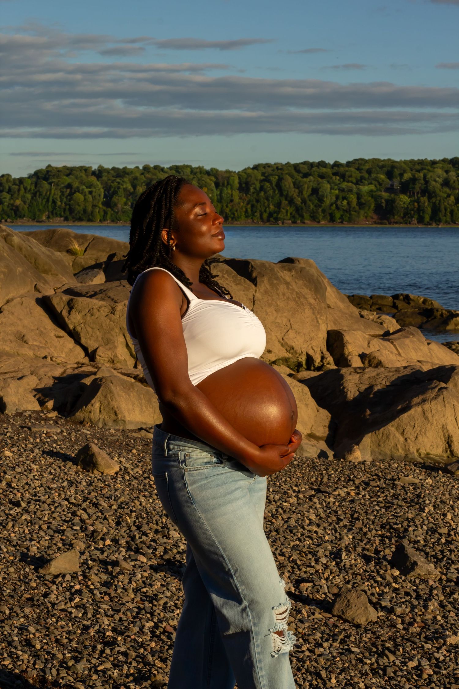 Pregnant woman in white top and jeans holds belly while standing on rocky beach at sunset.