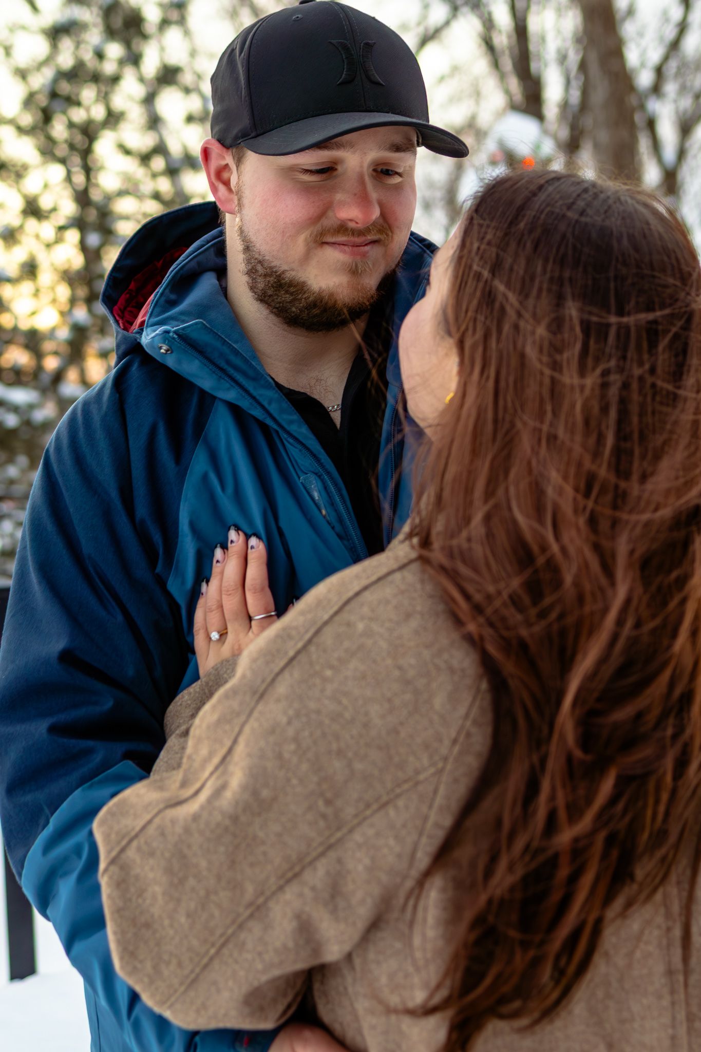 Romantic couple embracing outdoors in winter, man in black cap and blue jacket gazing lovingly at woman in tan coat.