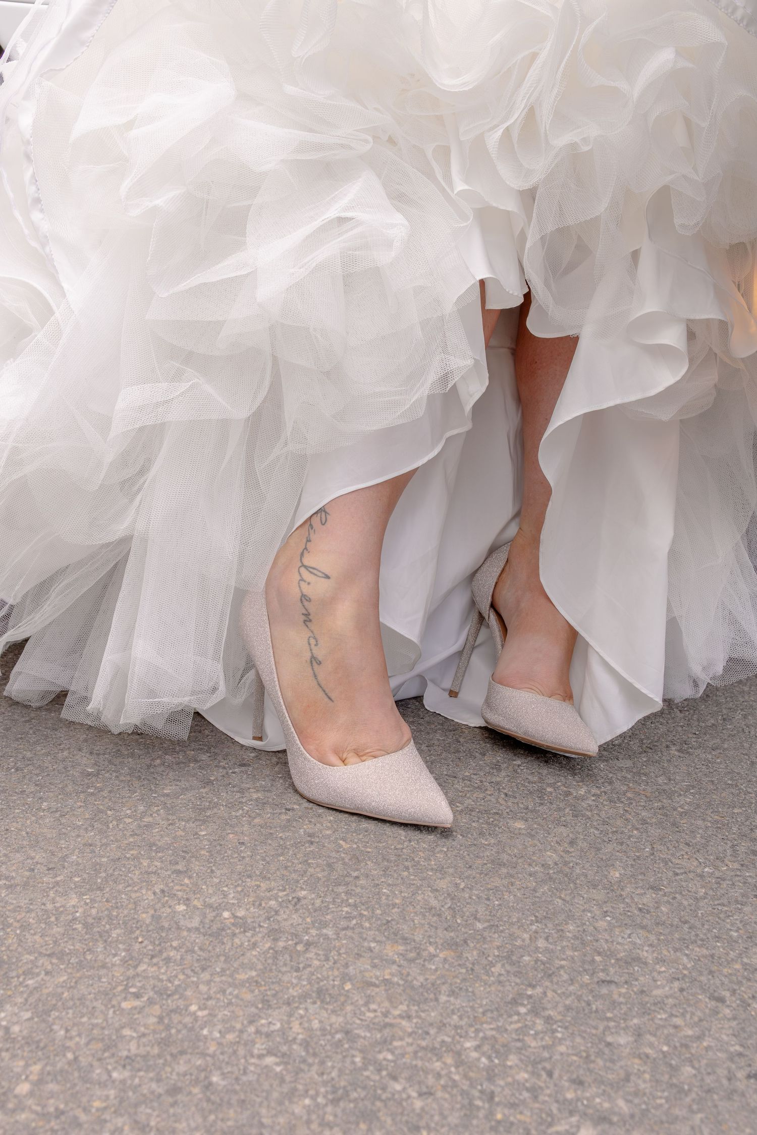 Bride wearing nude pointed-toe heels with a script tattoo on ankle, peeking beneath a ruffled white wedding gown.