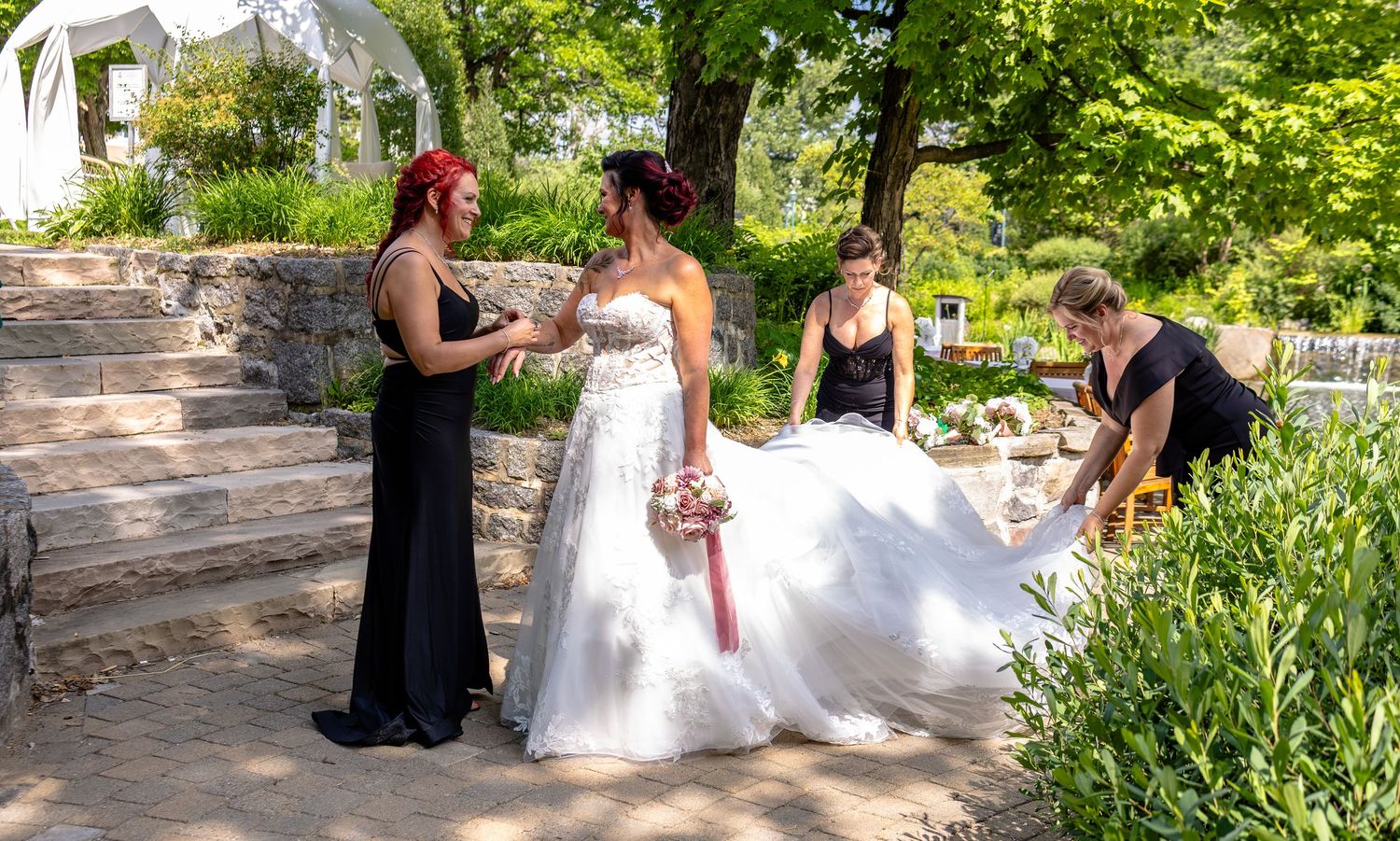 Bride in white gown with bridesmaids in black dresses adjusting her train in a sunny garden setting.
