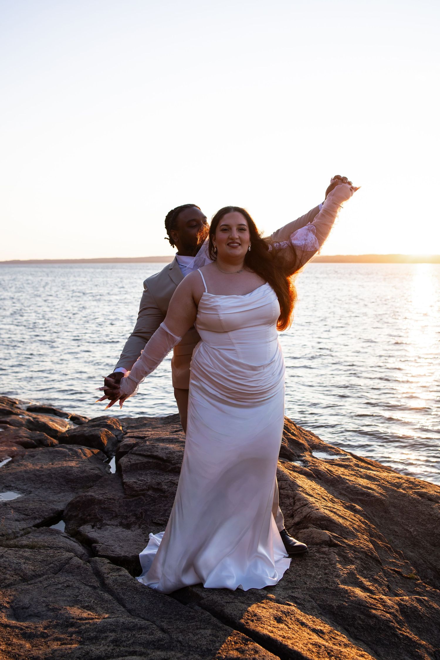 Joyful bride in white gown raises arm on rocky shore at sunset, groom in gray suit standing behind her by the water.
