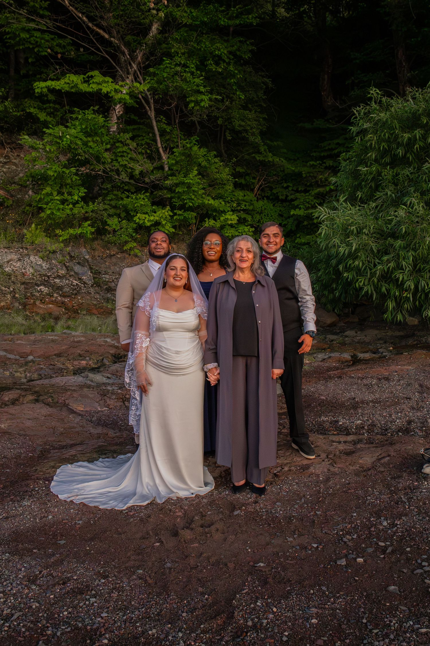Wedding party of five posing outdoors on rocky terrain with lush green forest backdrop, bride in white gown with train.