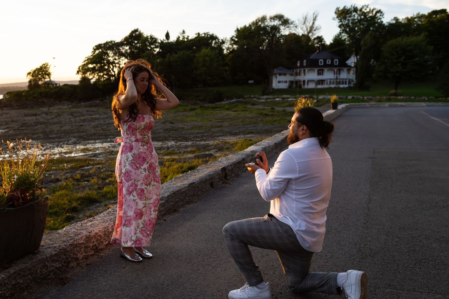 Man proposes on one knee to surprised woman in floral dress at sunset near waterfront estate.