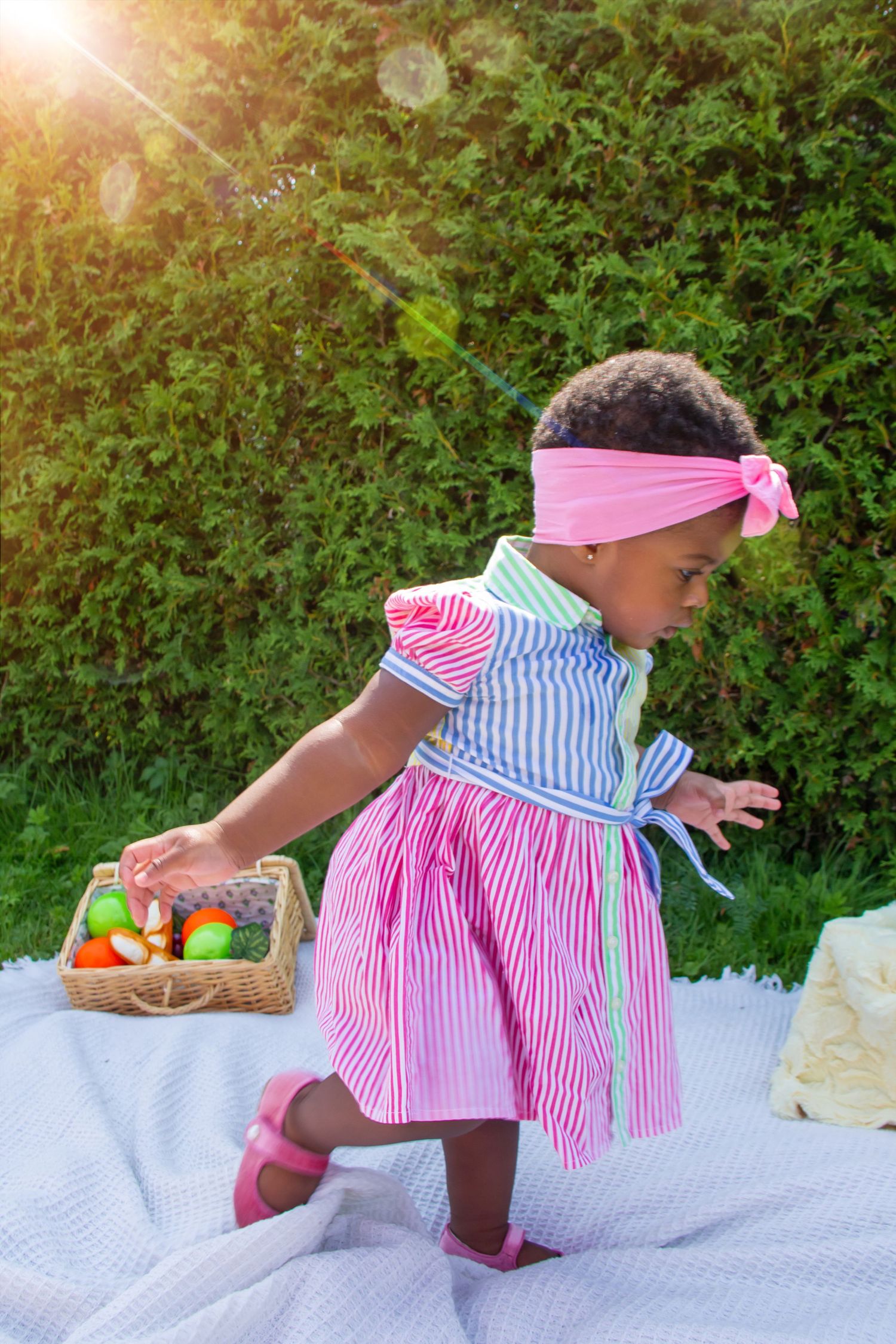 Adorable little girl in striped pink dress with bow headband reaching for Easter eggs in a wicker basket outdoors.