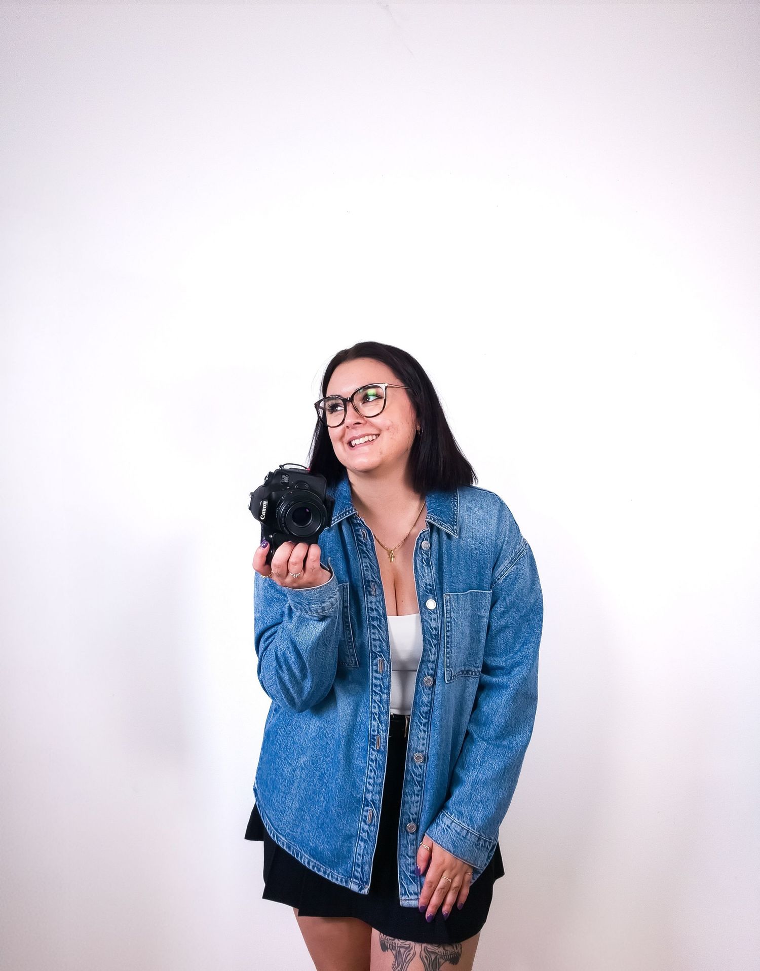 Smiling woman with glasses in denim jacket holding a Canon camera against a white background.