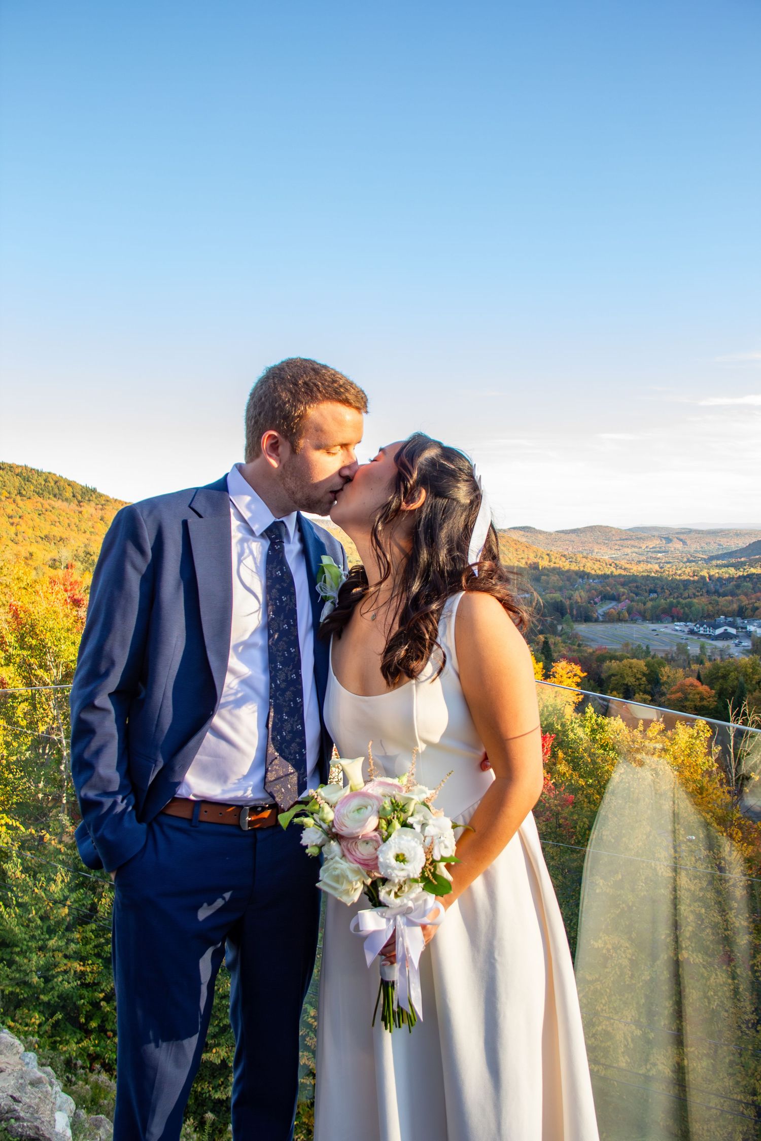 Bride and groom kissing on mountain overlook with colorful autumn foliage valley backdrop at sunset.
