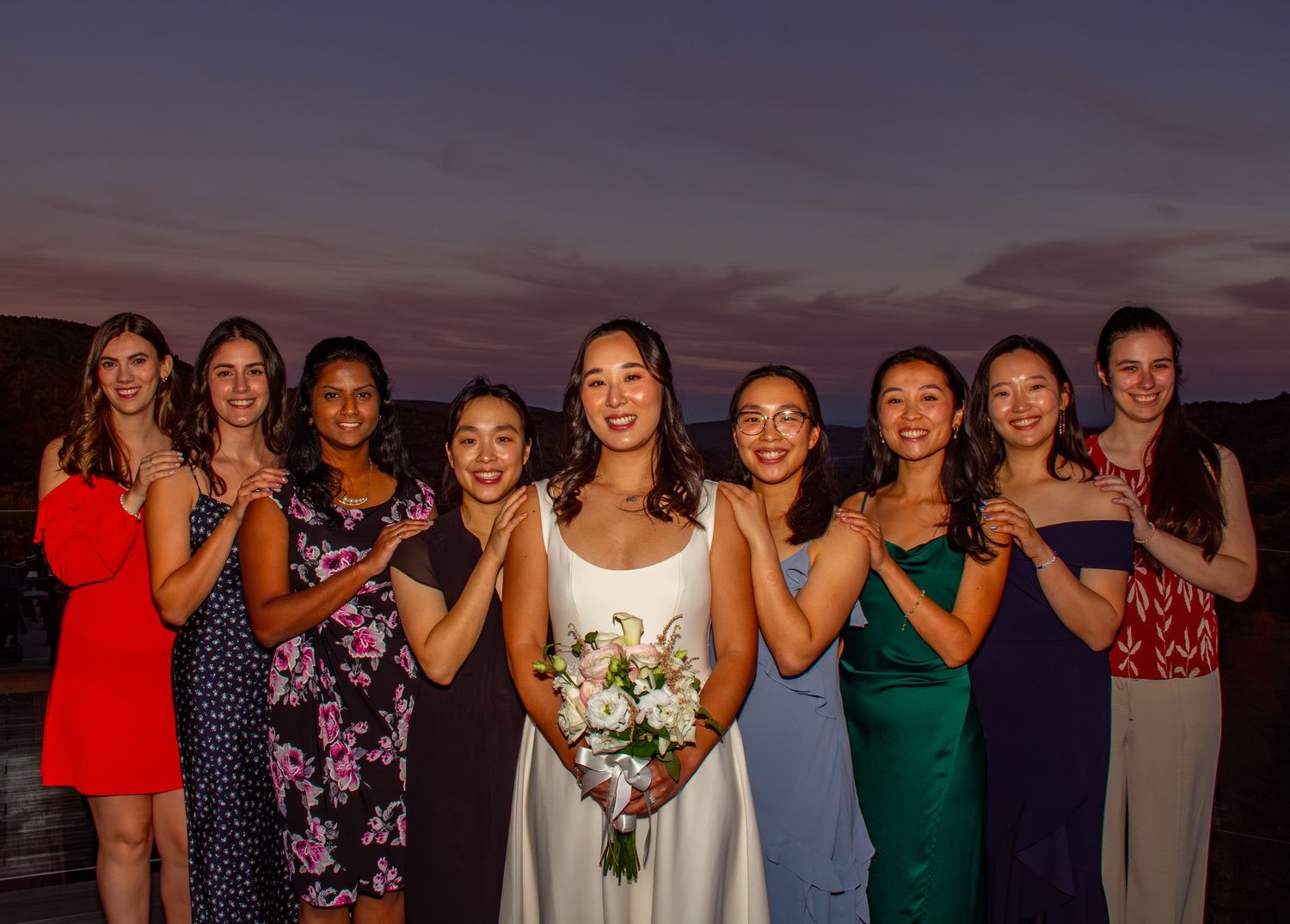 Bride in white dress poses with eight bridesmaids in colorful gowns at twilight outdoor wedding.