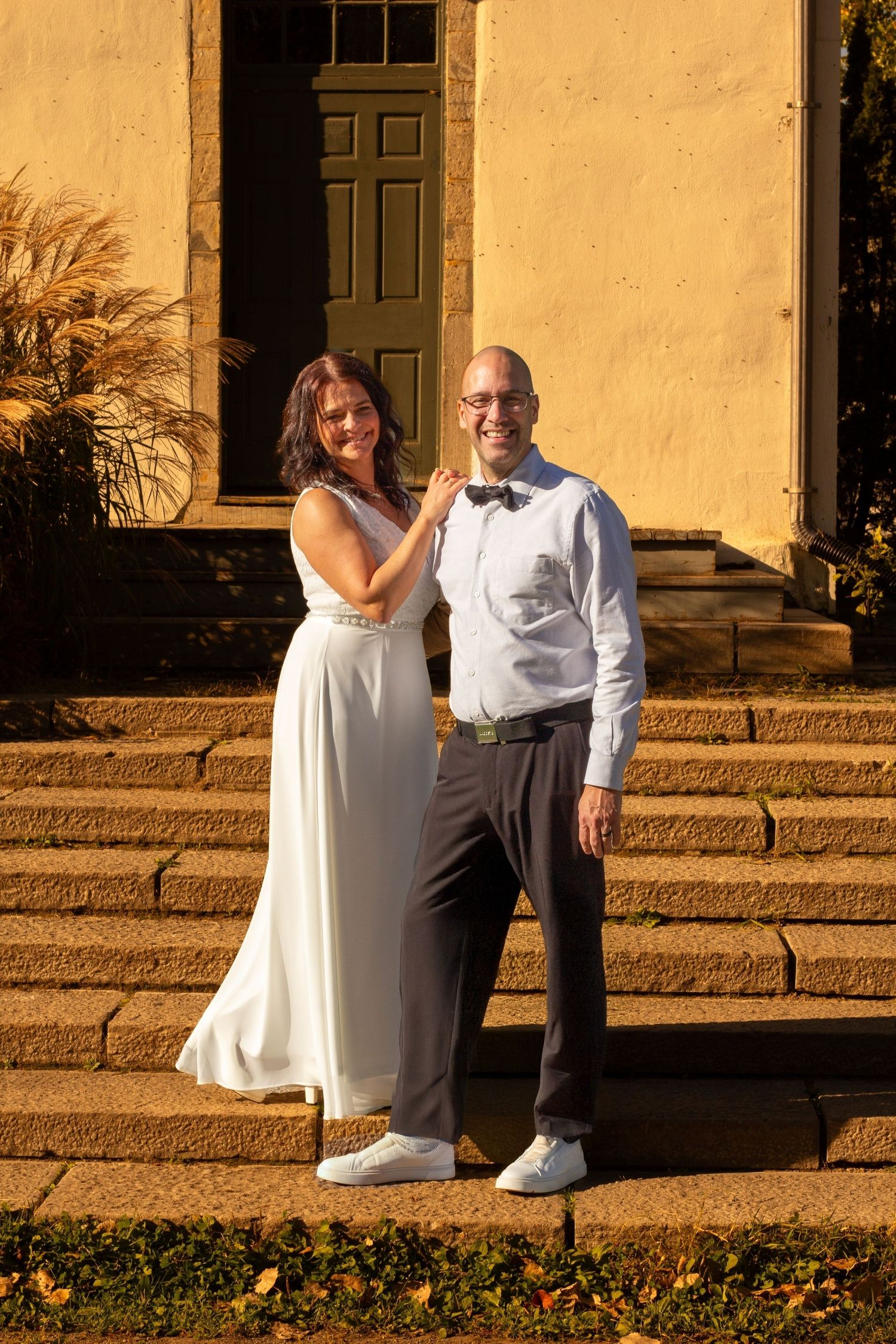 Smiling bride and bald groom pose together on stone steps of a golden sunlit historic building.