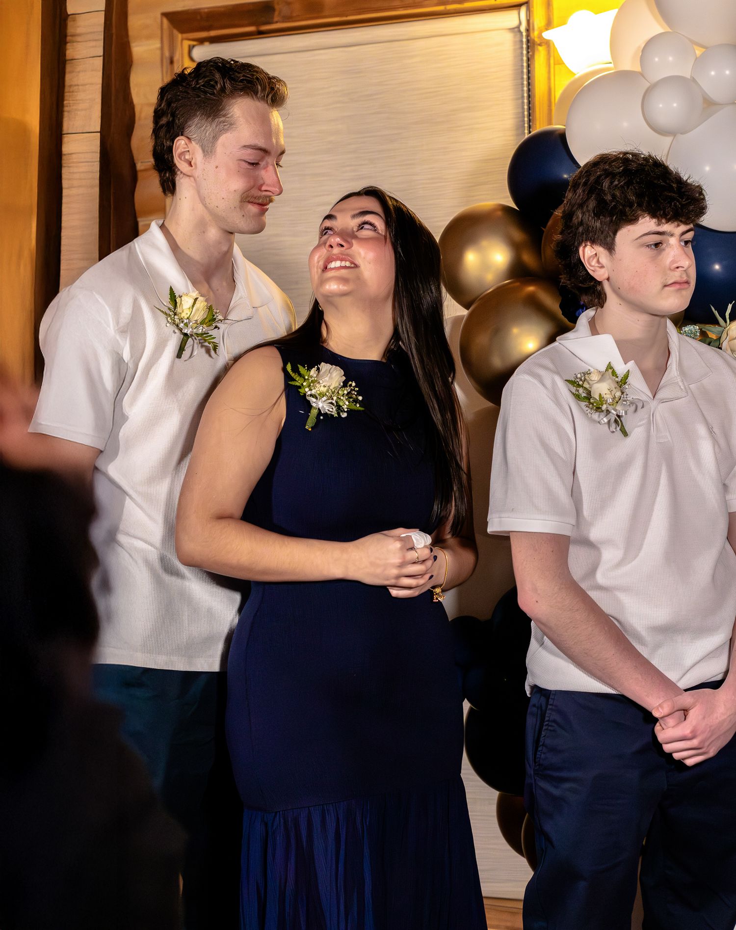 Three young wedding guests in navy and white attire with boutonnieres share emotional moment at decorated reception.