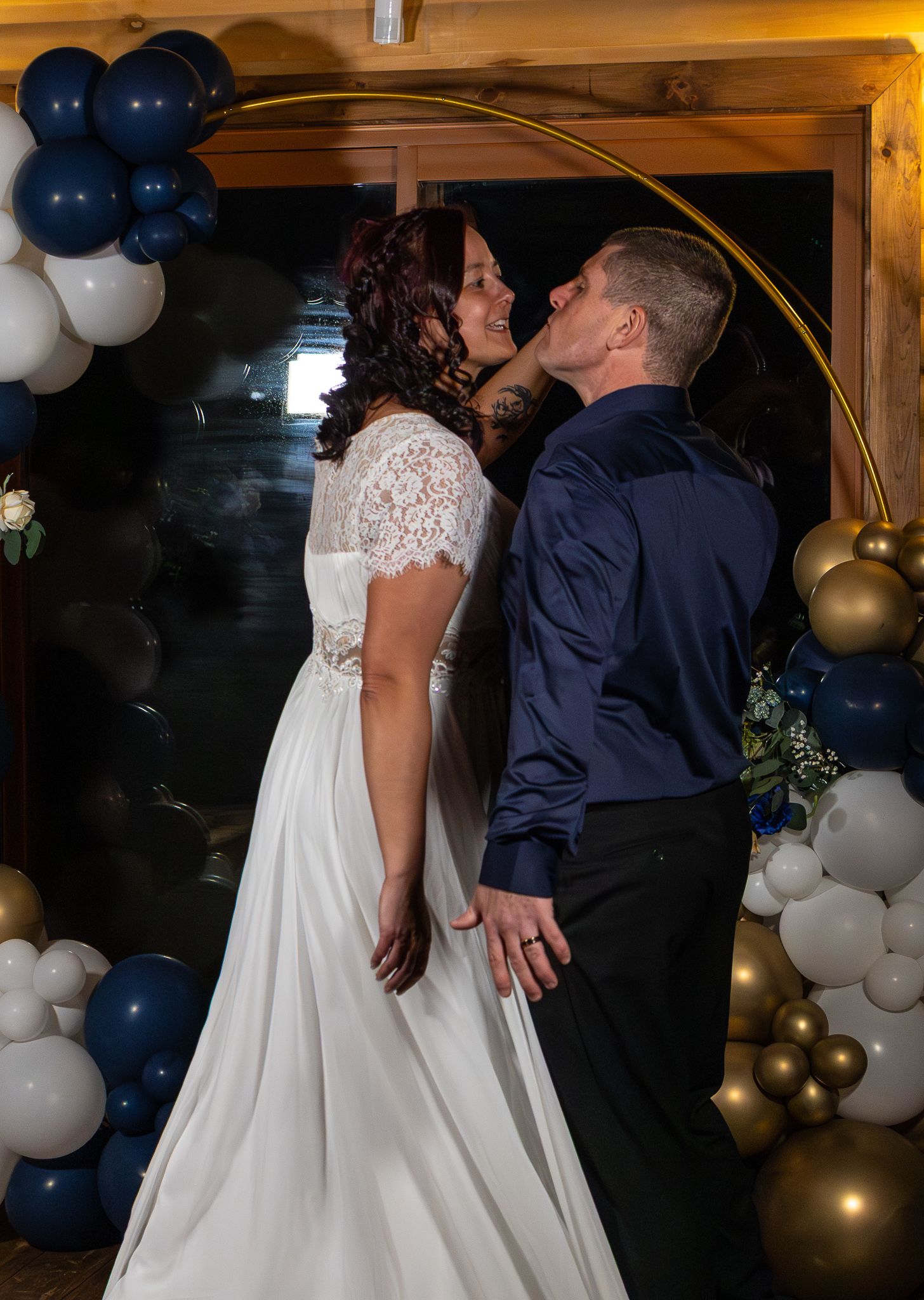 Bride and groom share a romantic kiss in front of a circular gold arch with navy and gold balloons.