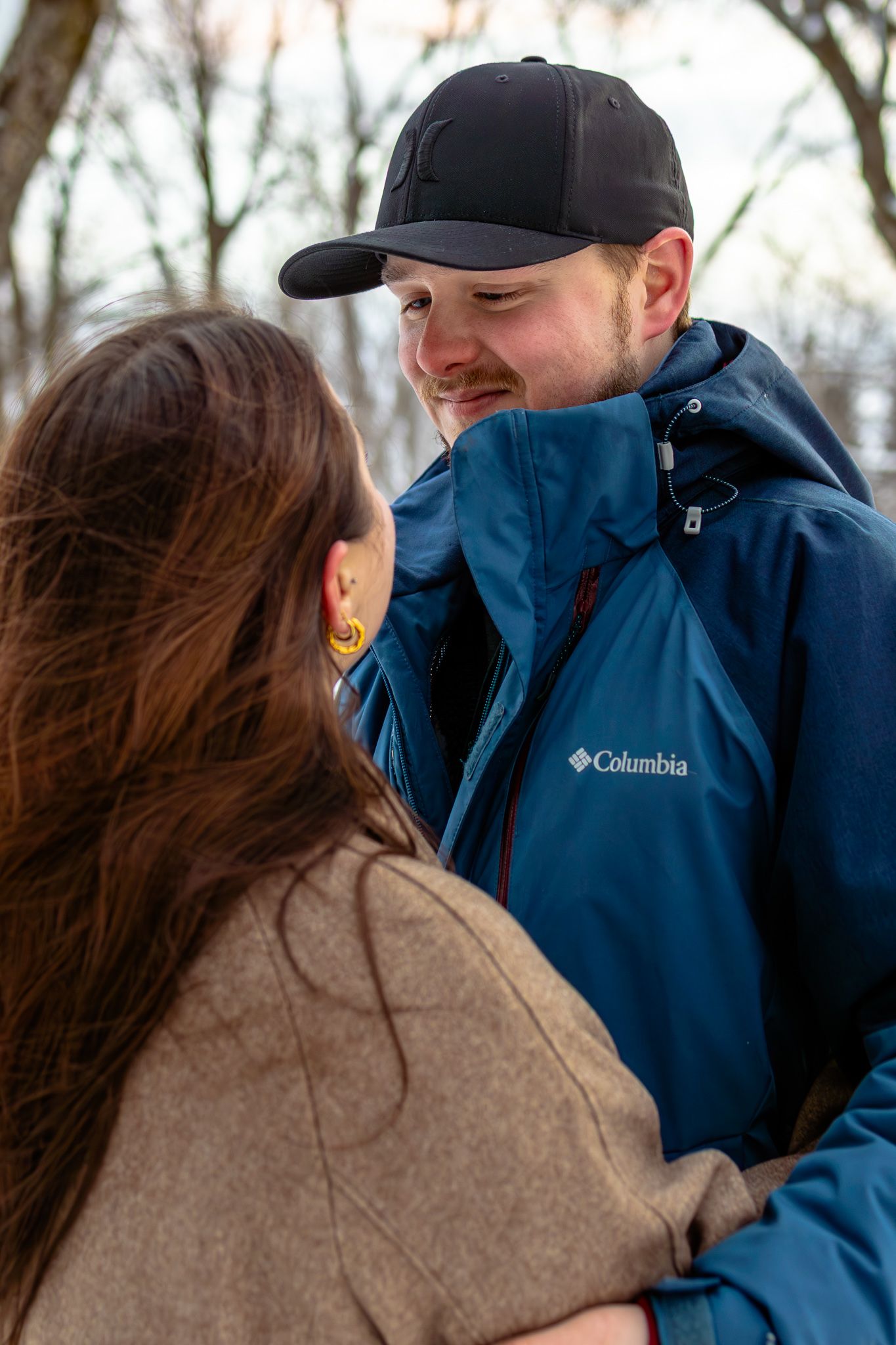 Man in Columbia blue jacket and black cap smiling warmly at woman with long brown hair in winter outdoor setting.
