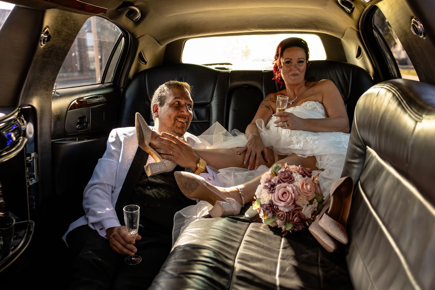 Bride and groom in wedding attire sitting in a luxury limousine, holding champagne glasses and a floral bouquet.