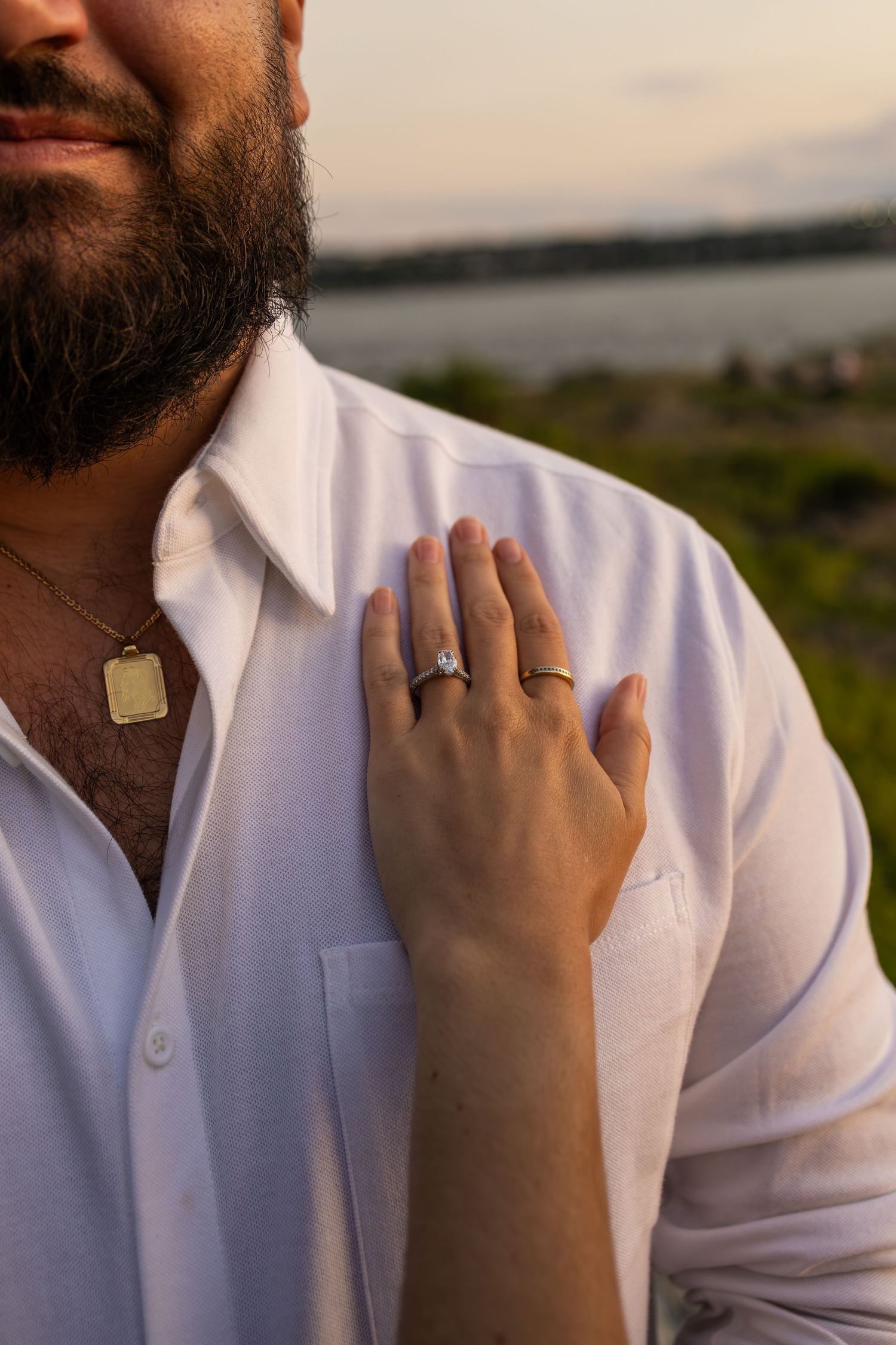 Close-up of a woman's hand with an engagement ring resting on a bearded man's white linen shirt outdoors.