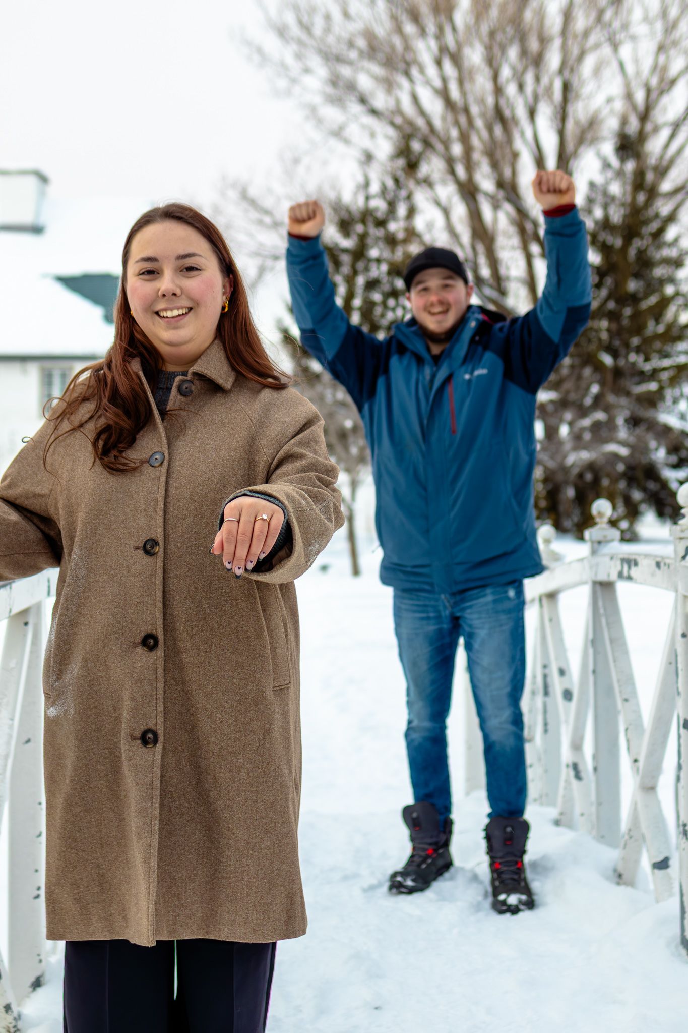 Excited newly engaged couple celebrating in snow, woman showing engagement ring while man celebrates with raised fists behind her.