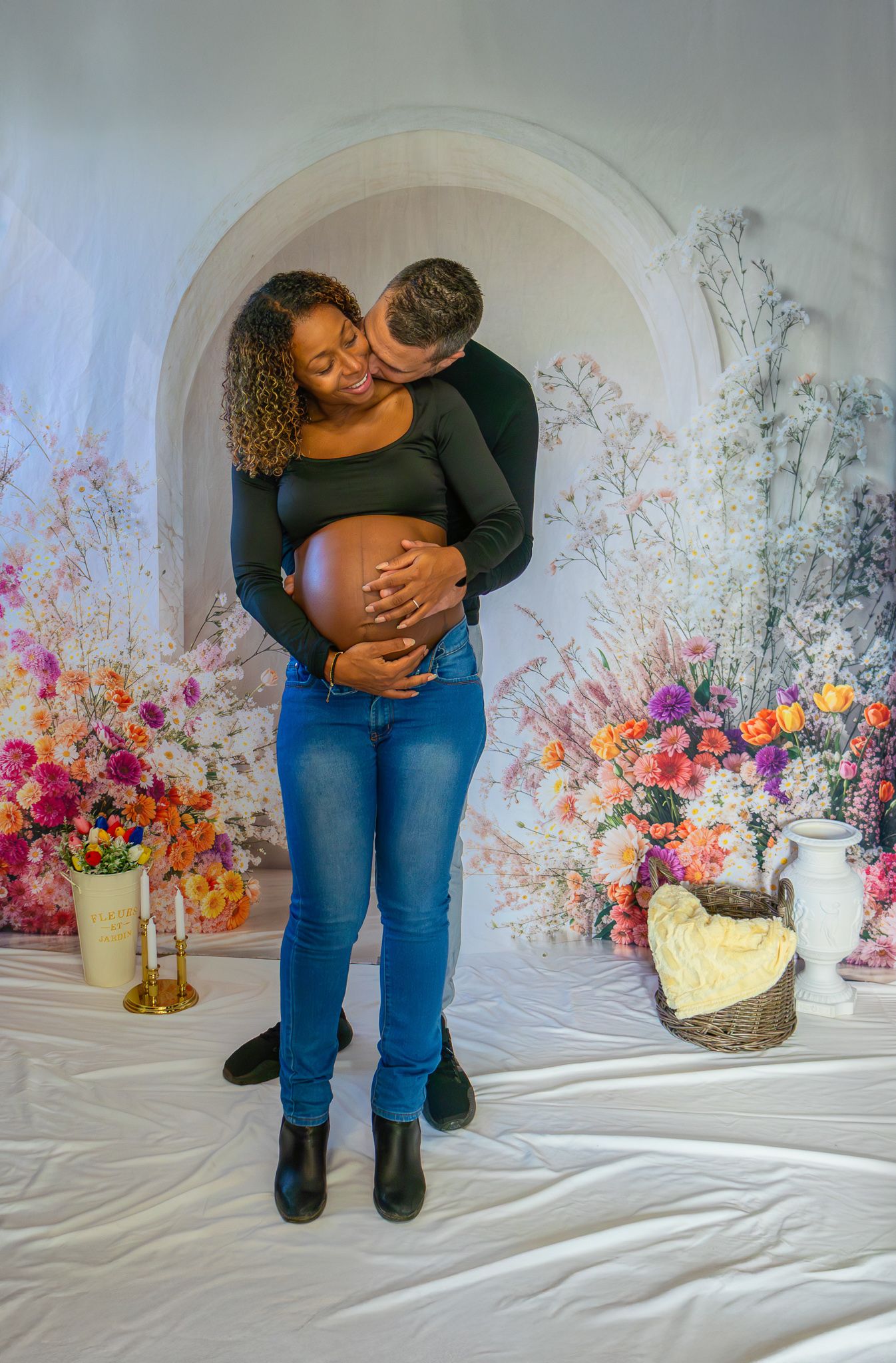 Couple sharing intimate embrace during maternity photoshoot with colorful floral arrangement background.