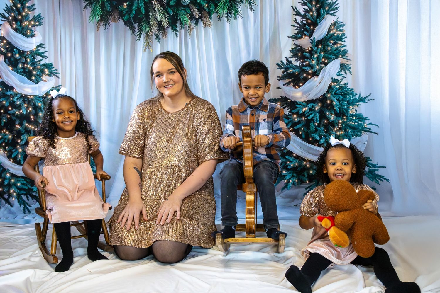 Woman in gold sequin dress posing with three children in front of decorated Christmas trees.