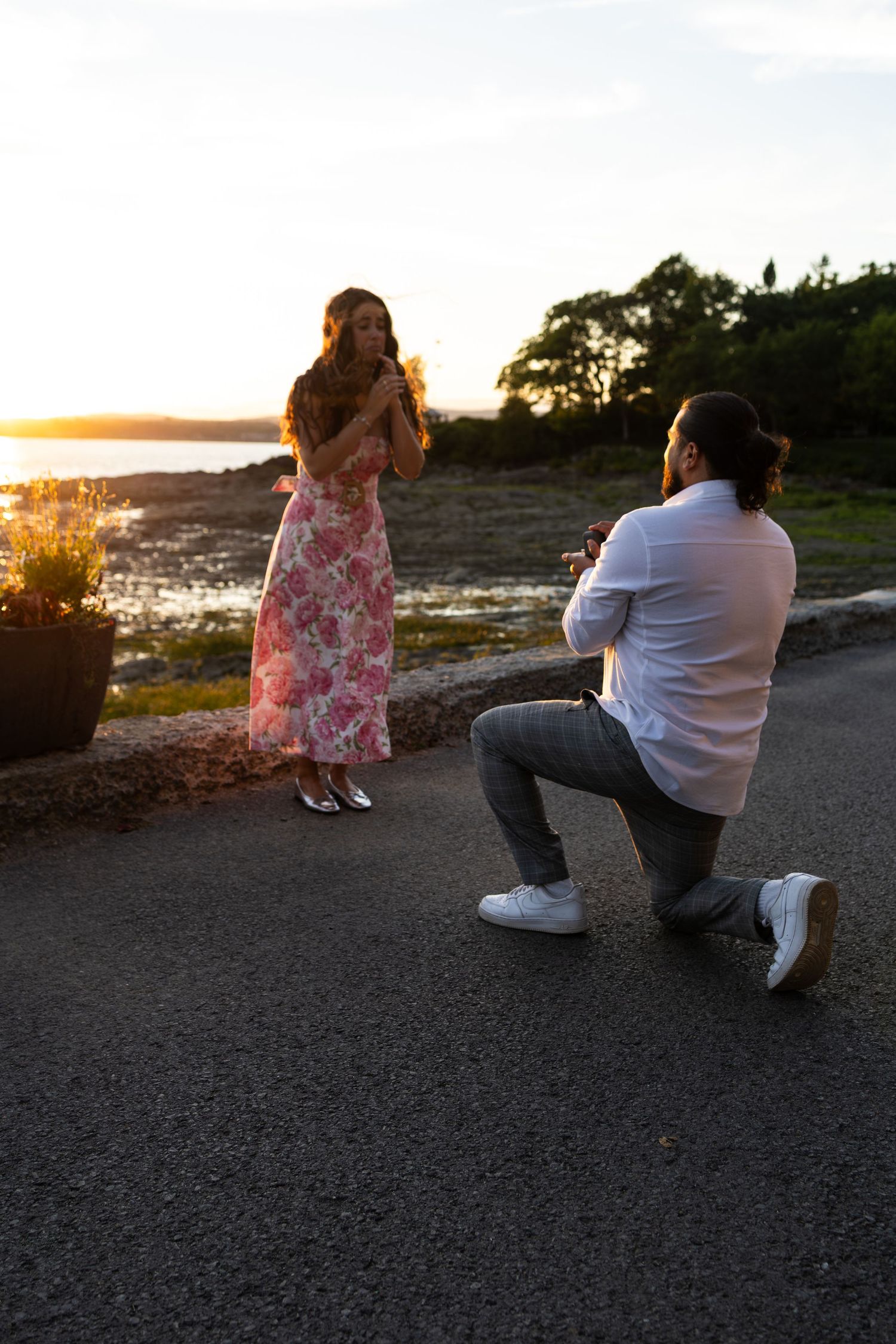 Man proposing on one knee to woman in floral dress at sunset by the water.