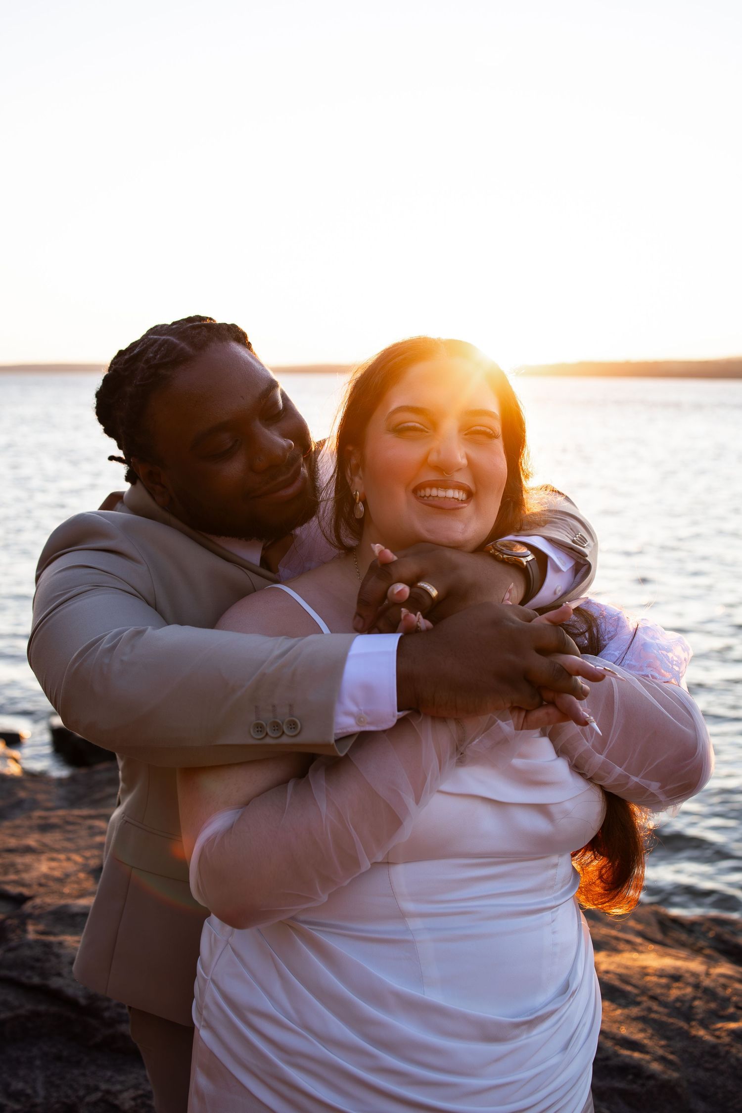 Couple embracing at sunset by the water, groom in tan suit hugging bride in white dress, golden backlight.
