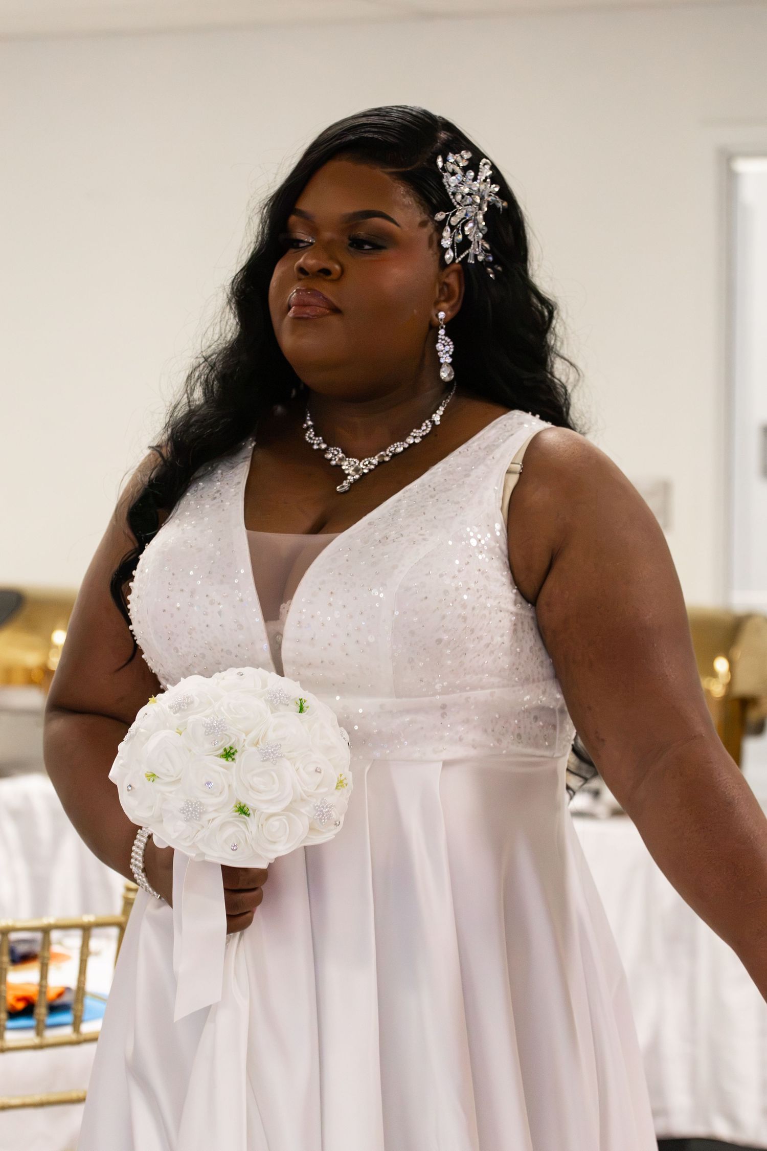 A plus-size bride in a white sequined gown holds a white bouquet, wearing diamond jewelry and a floral hair accessory.
