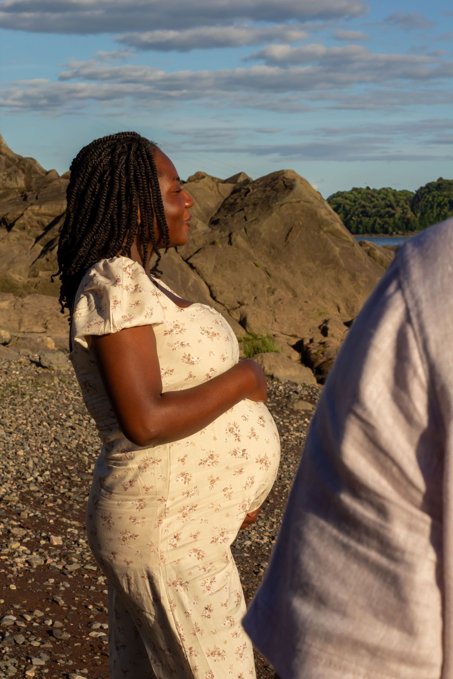 Pregnant Black woman in floral dress cradling baby bump on rocky shoreline at golden hour.