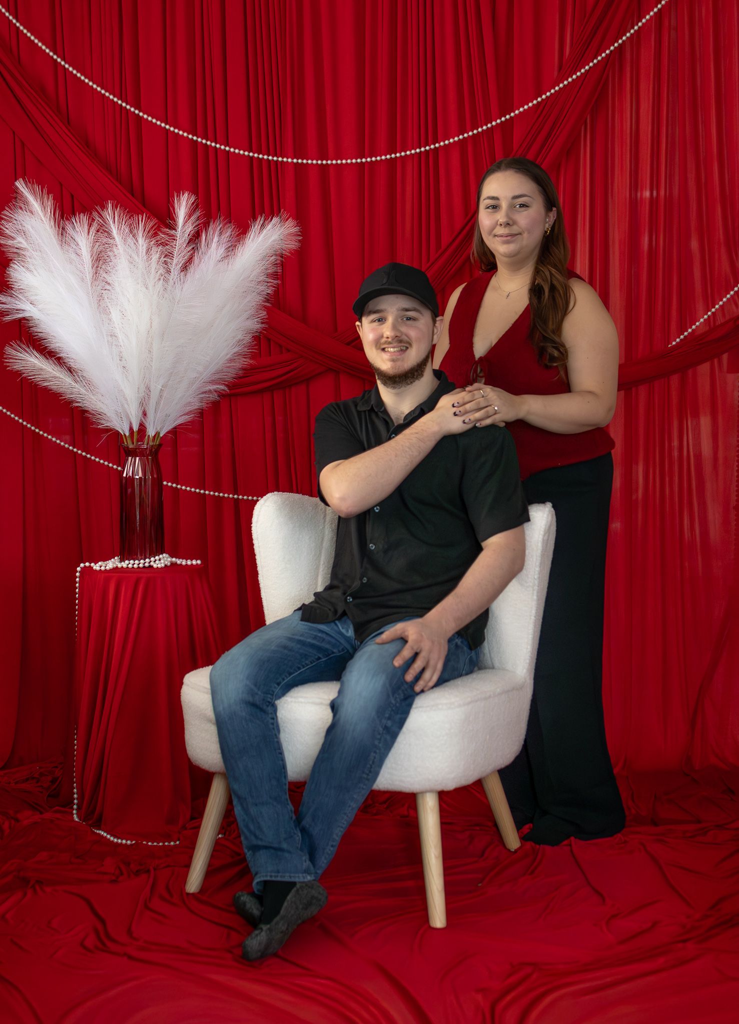 Couple posing with man seated in white chair and woman standing beside pampas grass decor.