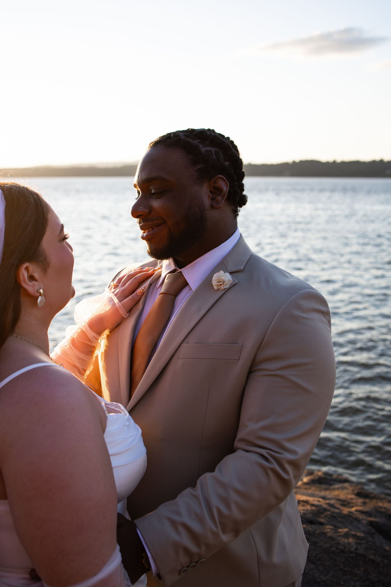 A bride and groom share a tender moment by the water at sunset, with the groom in a tan suit and pink tie.